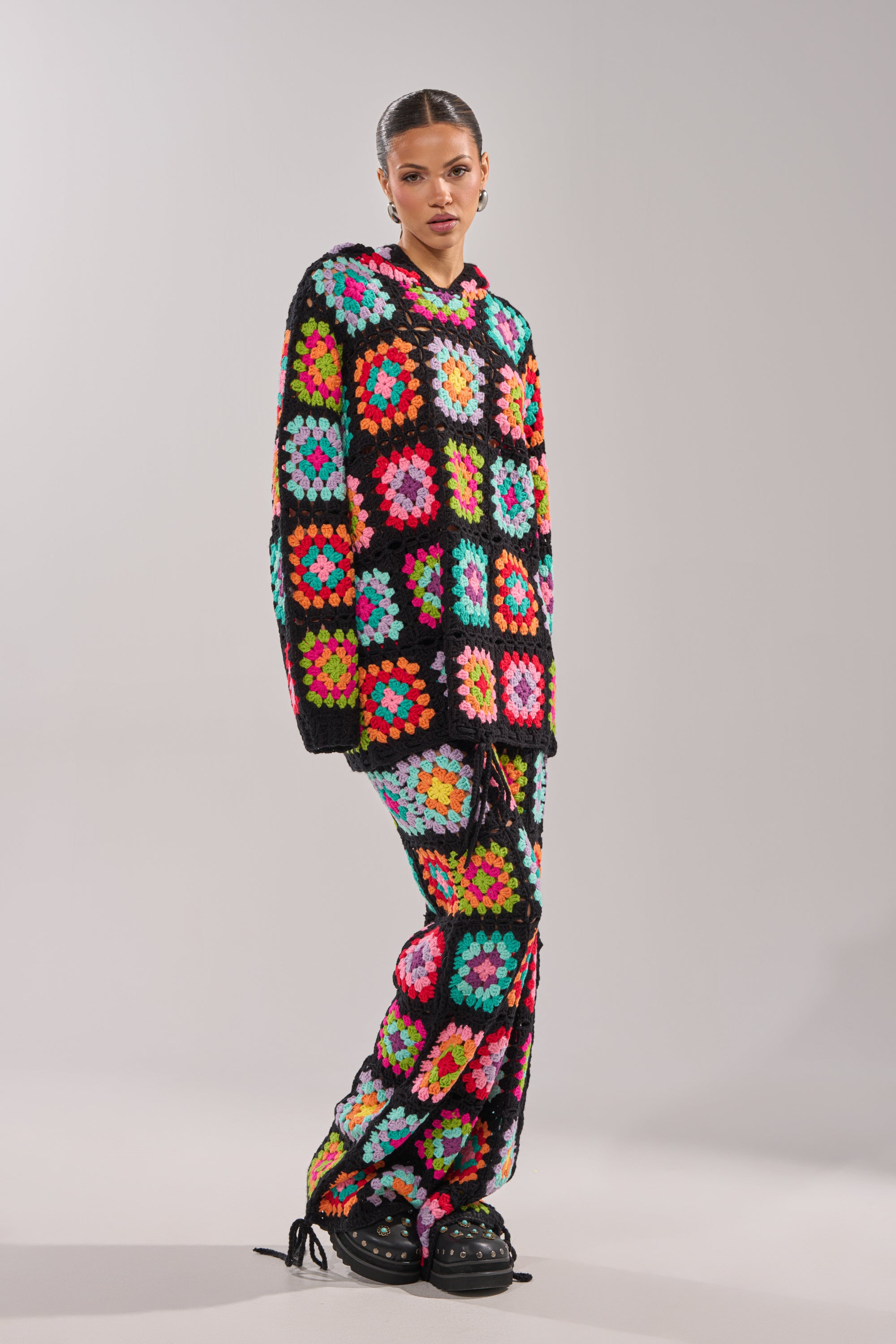 A woman stands against a plain background wearing the FESTIVAL GIRLY OVERSIZED CROCHET SWEATSHIRT IN MULTI, featuring colorful granny-square patchwork, paired with matching pants and black sandals.