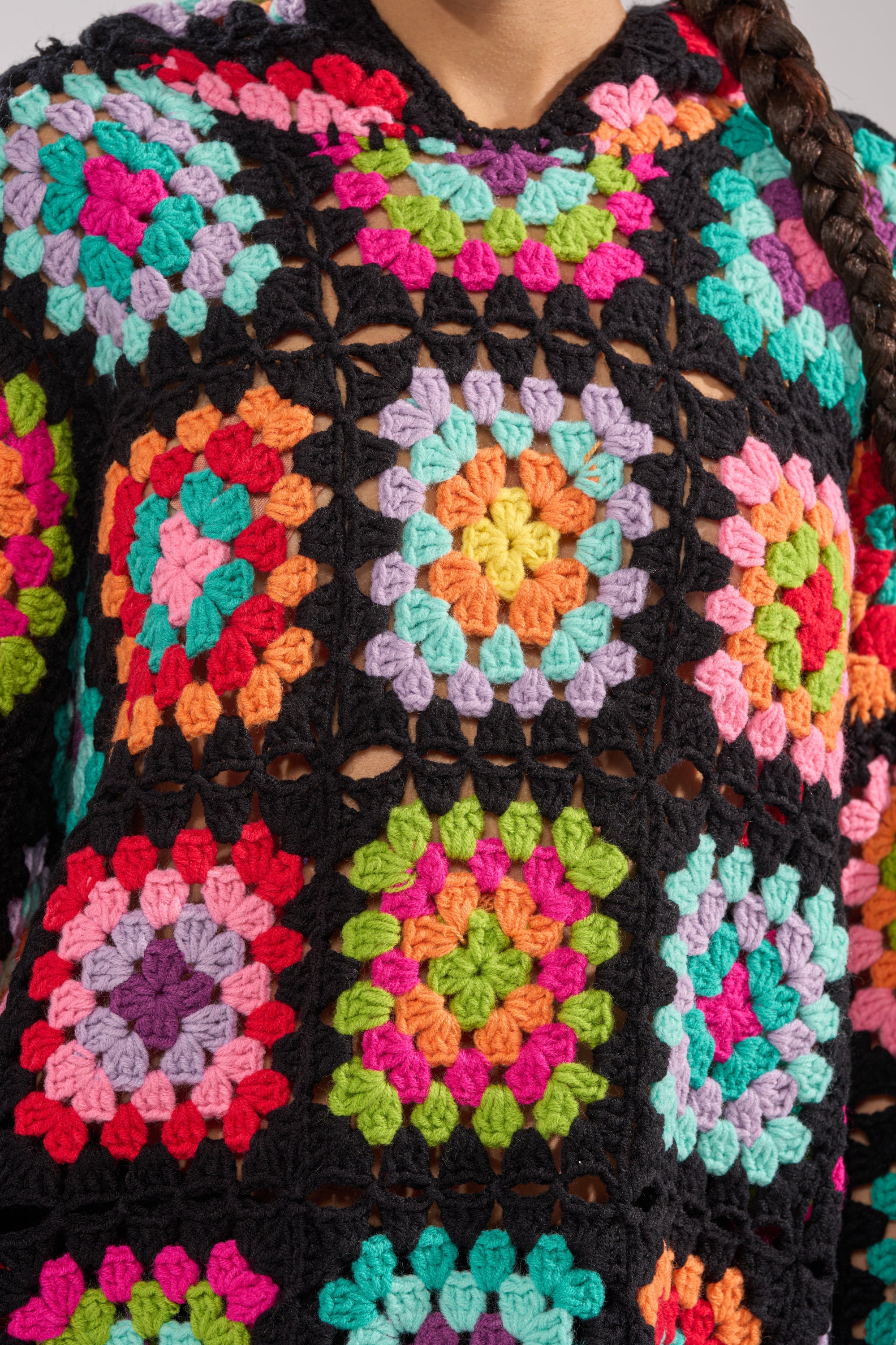 Close-up of the FESTIVAL GIRLY OVERSIZED CROCHET SWEATSHIRT IN MULTI, featuring colorful granny-square patchwork in pink, green, orange, yellow, purple, and turquoise on black. A braid drapes over the shoulder.