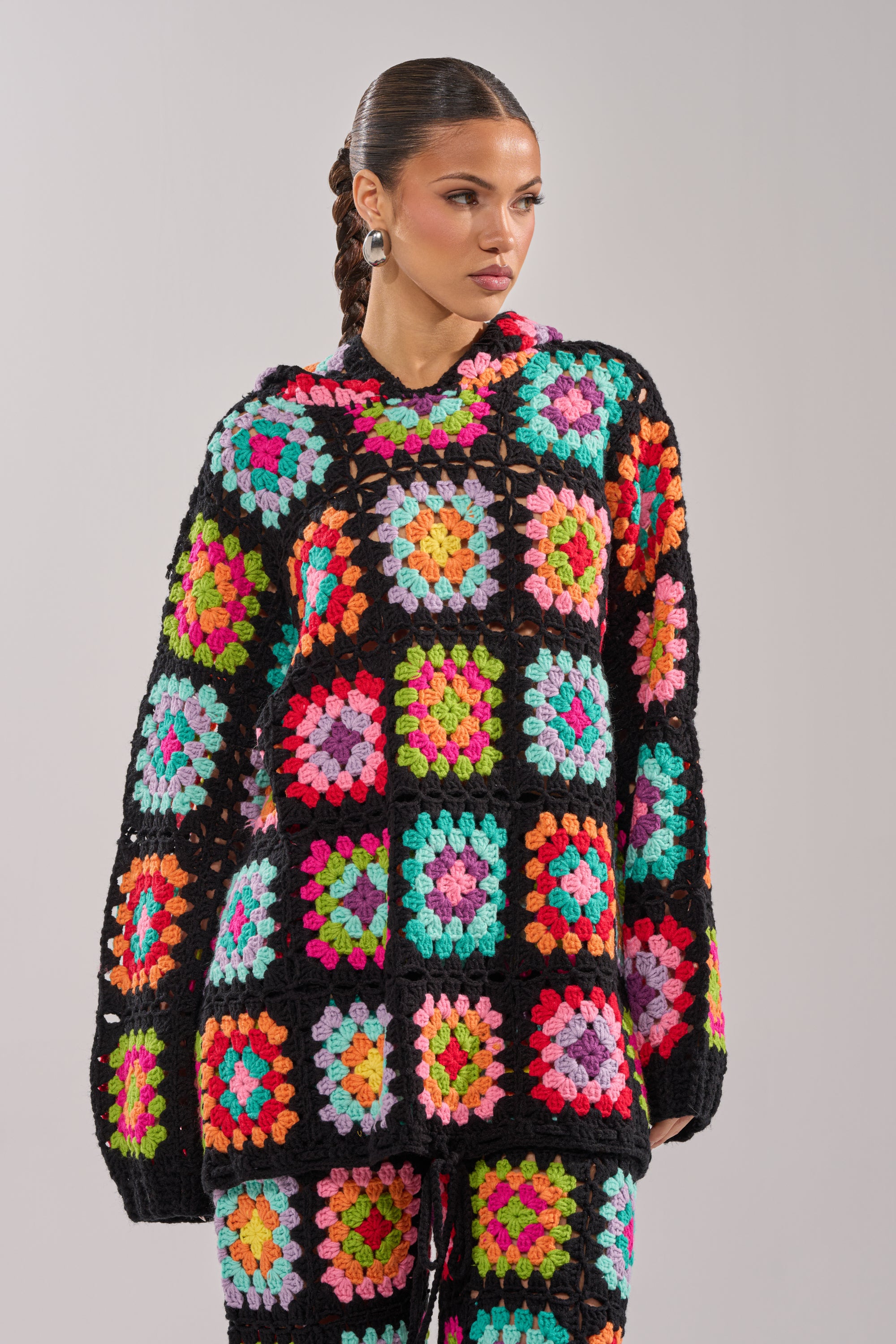 A woman wears the FESTIVAL GIRLY OVERSIZED CROCHET SWEATSHIRT IN MULTI, featuring a colorful patchwork design with a black base and multicolored squares, as she stands against a plain grey background looking to the side.