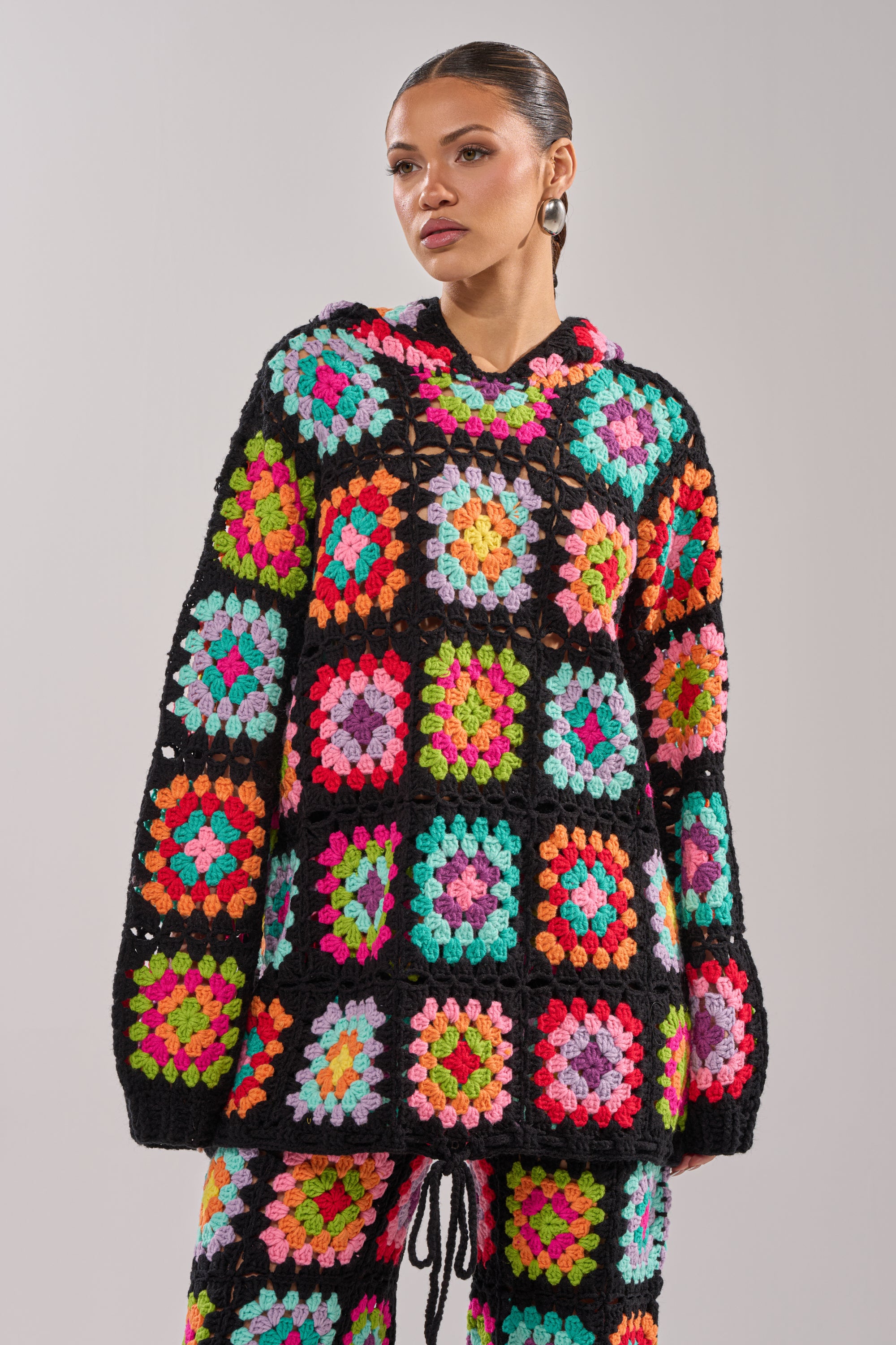 A woman models the FESTIVAL GIRLY OVERSIZED CROCHET SWEATSHIRT IN MULTI, featuring vibrant granny-square patchwork on black. She pairs it with matching pants and stands against a light gray background, gazing thoughtfully to the side.