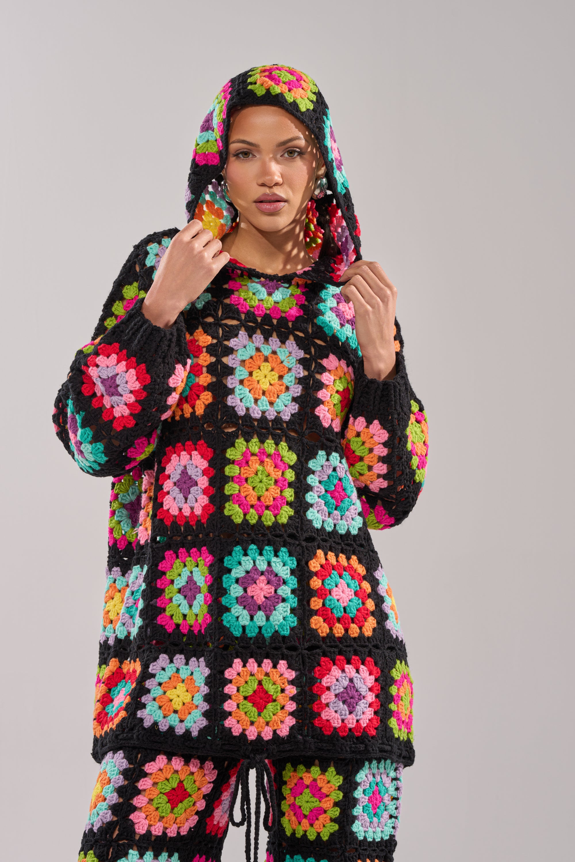 A woman wears the FESTIVAL GIRLY OVERSIZED CROCHET SWEATSHIRT IN MULTI with matching pants, showcasing colorful granny-square patchwork on black, as she stands before a plain light grey background.