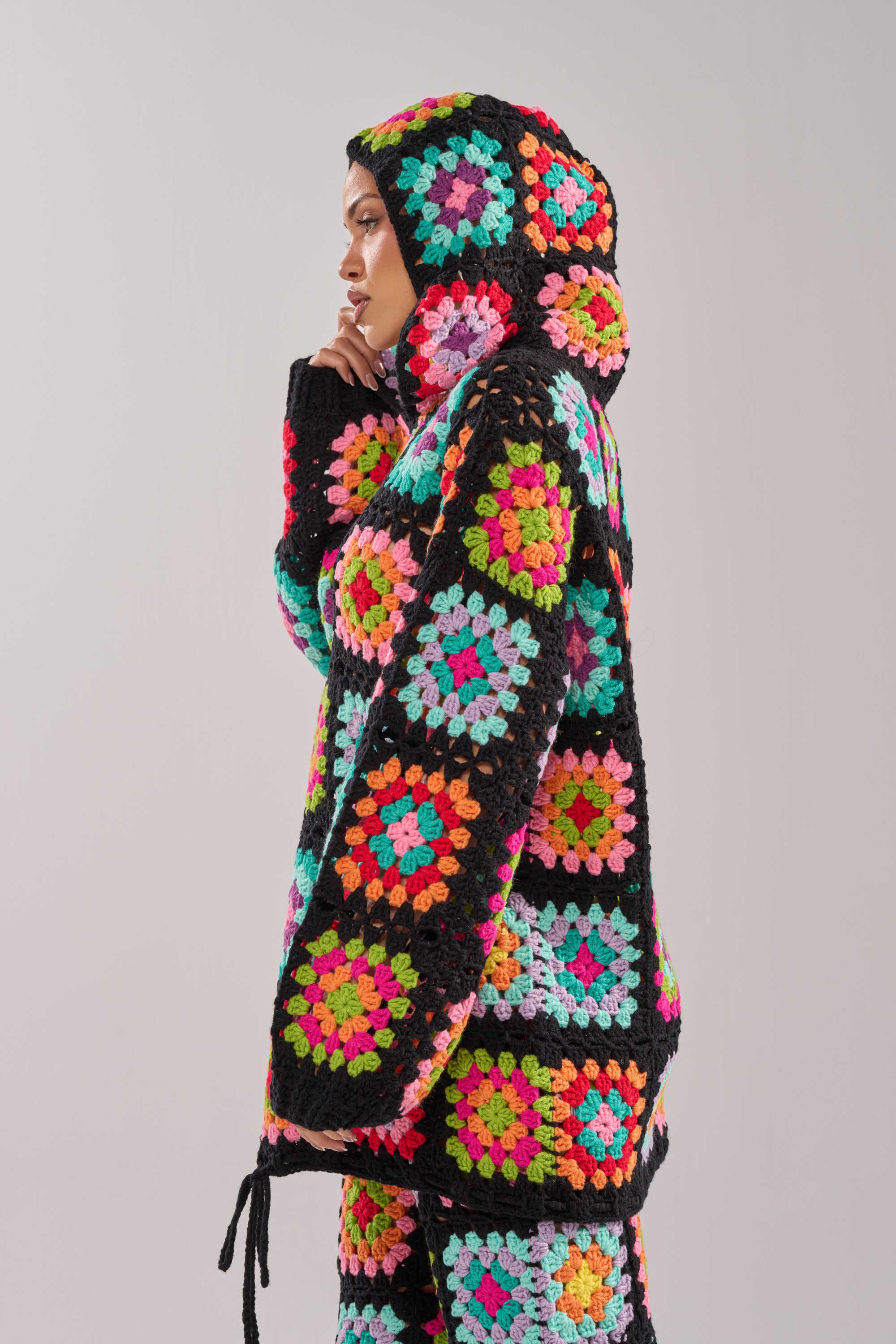 A person stands in profile against a plain background, wearing the FESTIVAL GIRLY OVERSIZED CROCHET SWEATSHIRT IN MULTI—an oversized, colorful granny-square hoodie with black borders and a hood pulled over their head.