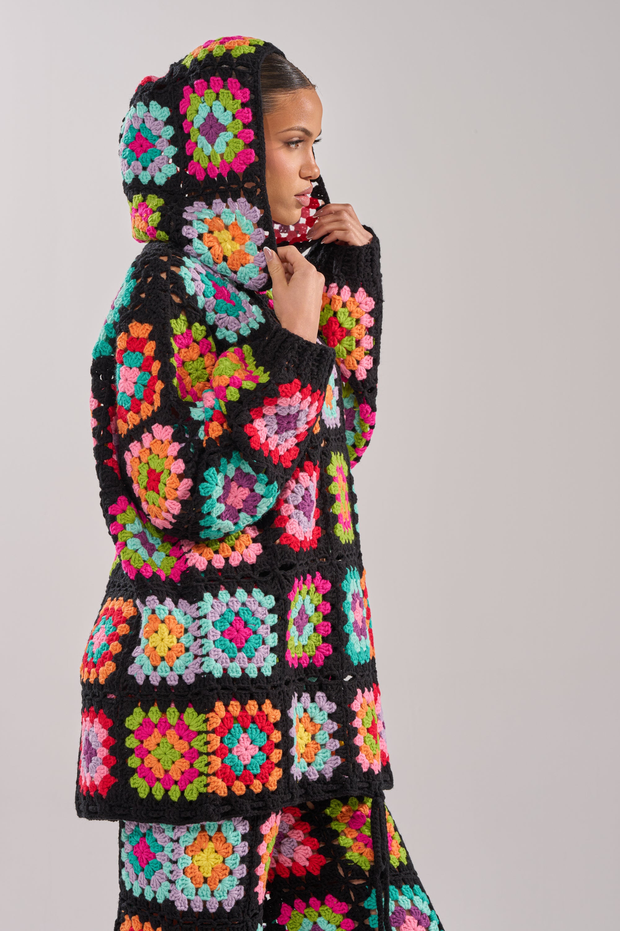 Wearing the FESTIVAL GIRLY OVERSIZED CROCHET SWEATSHIRT IN MULTI, featuring bright granny-square patchwork bordered in black, a person stands against a plain light gray background.