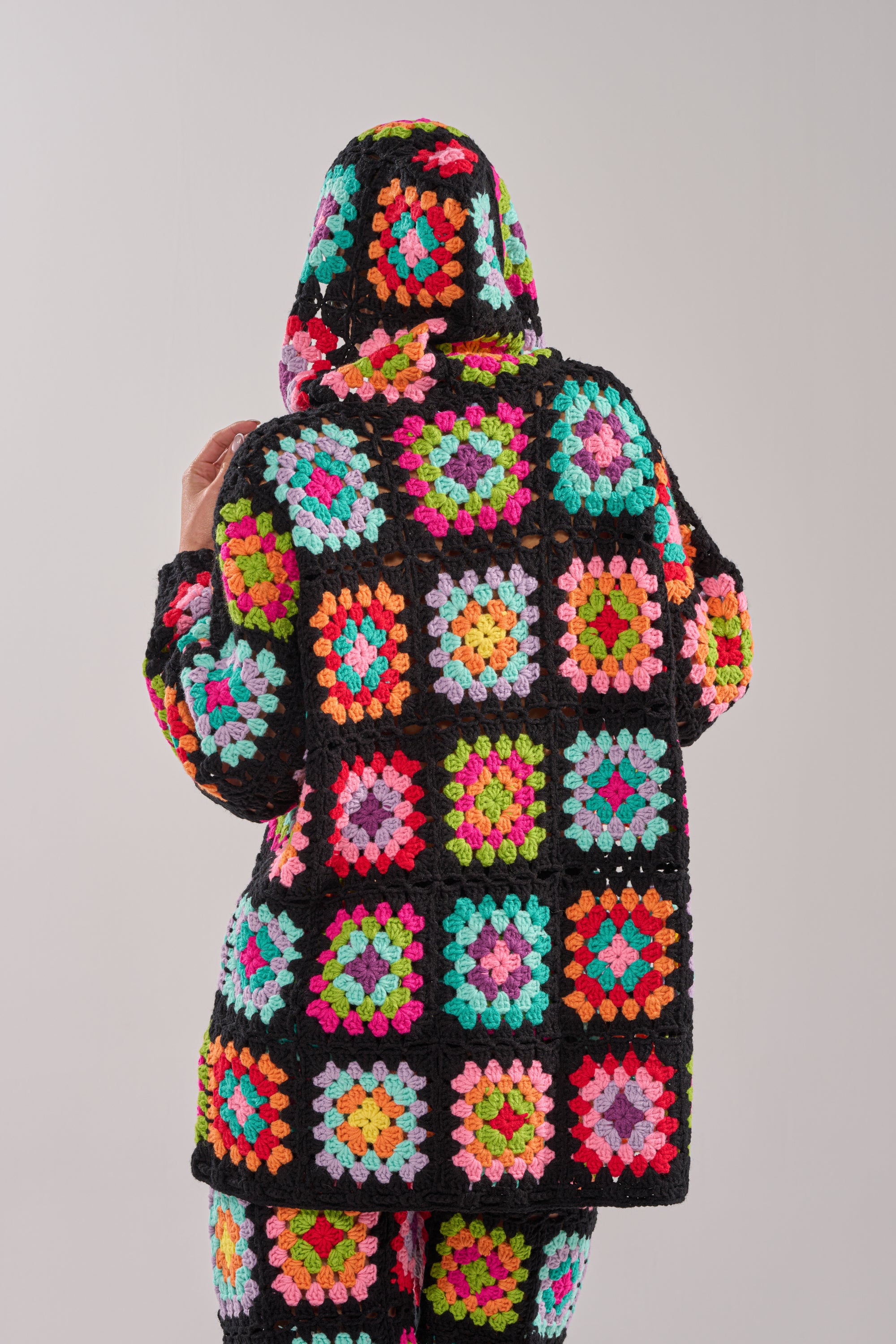 A person wearing the FESTIVAL GIRLY OVERSIZED CROCHET SWEATSHIRT IN MULTI and matching pants with a granny-square patchwork pattern faces away from the camera against a plain background.