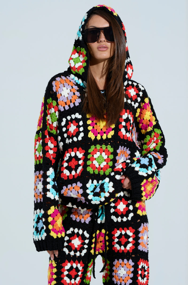 Modeling large black sunglasses, a person showcases the FESTIVAL GIRLY OVERSIZED CROCHET SWEATSHIRT from AKIRA Label. Featuring multicolored square patterns on a black backdrop, it creates an eye-catching silhouette against the light gray background.