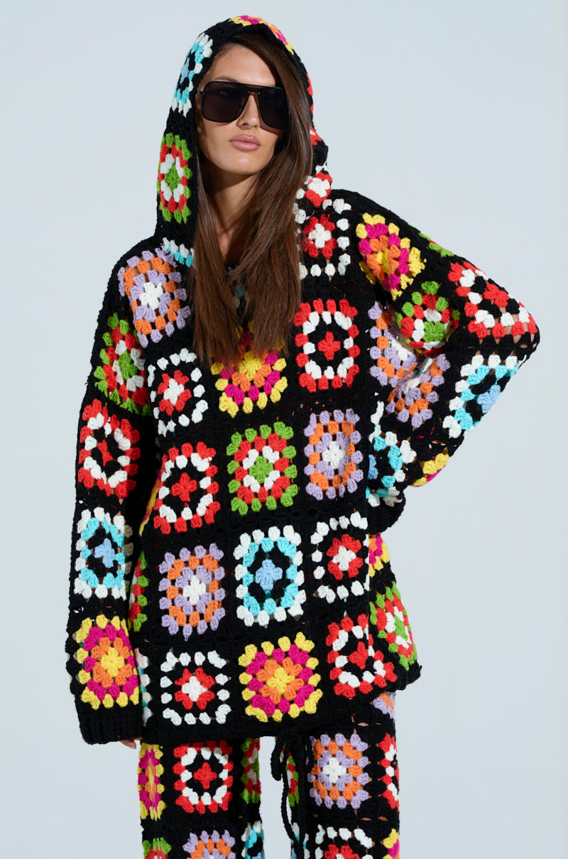 A person confidently poses against a plain background wearing an eye-catching, colorful crochet outfit with a relaxed oversized silhouette. The ensemble includes the FESTIVAL GIRLY OVERSIZED CROCHET SWEATSHIRT and is adorned with patchwork multicolored floral patterns. They have long hair and large black sunglasses.