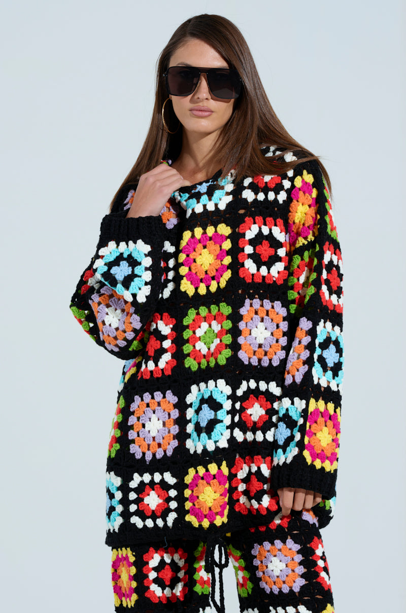 Wearing a Festival Girly Oversized Crochet Sweatshirt crafted from vibrant crocheted granny squares in black, red, blue, yellow, and pink, the person complements their long hair and large black sunglasses against a simple light background.