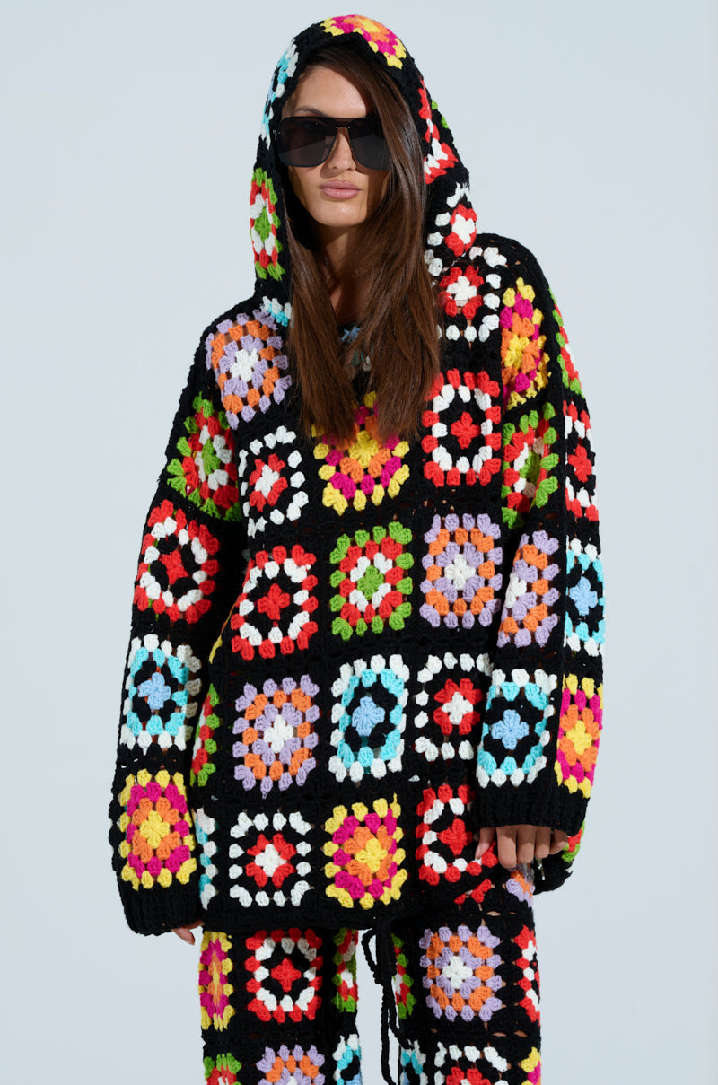 Wearing sunglasses and a FESTIVAL GIRLY OVERSIZED CROCHET SWEATSHIRT, the outfit has a relaxed silhouette with colorful crochet knit squares on black, creating a festive vibe. Paired with matching pants, it's a truly girly ensemble.