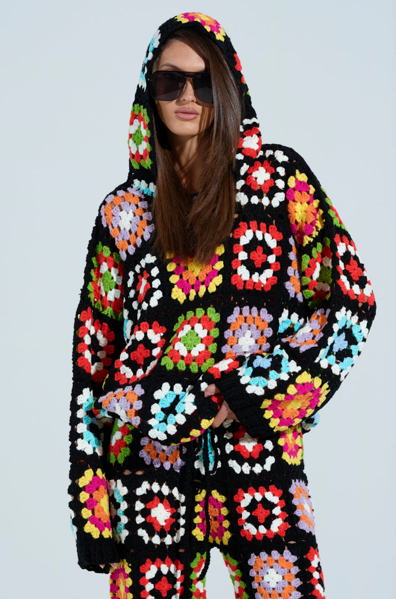 A model in sunglasses showcases the AKIRA Label FESTIVAL GIRLY OVERSIZED CROCHET SWEATSHIRT and pants, adorned with vibrant squares in red, green, blue, pink, and yellow against black. The colorful sweatshirt with hoodie pops against the light backdrop.