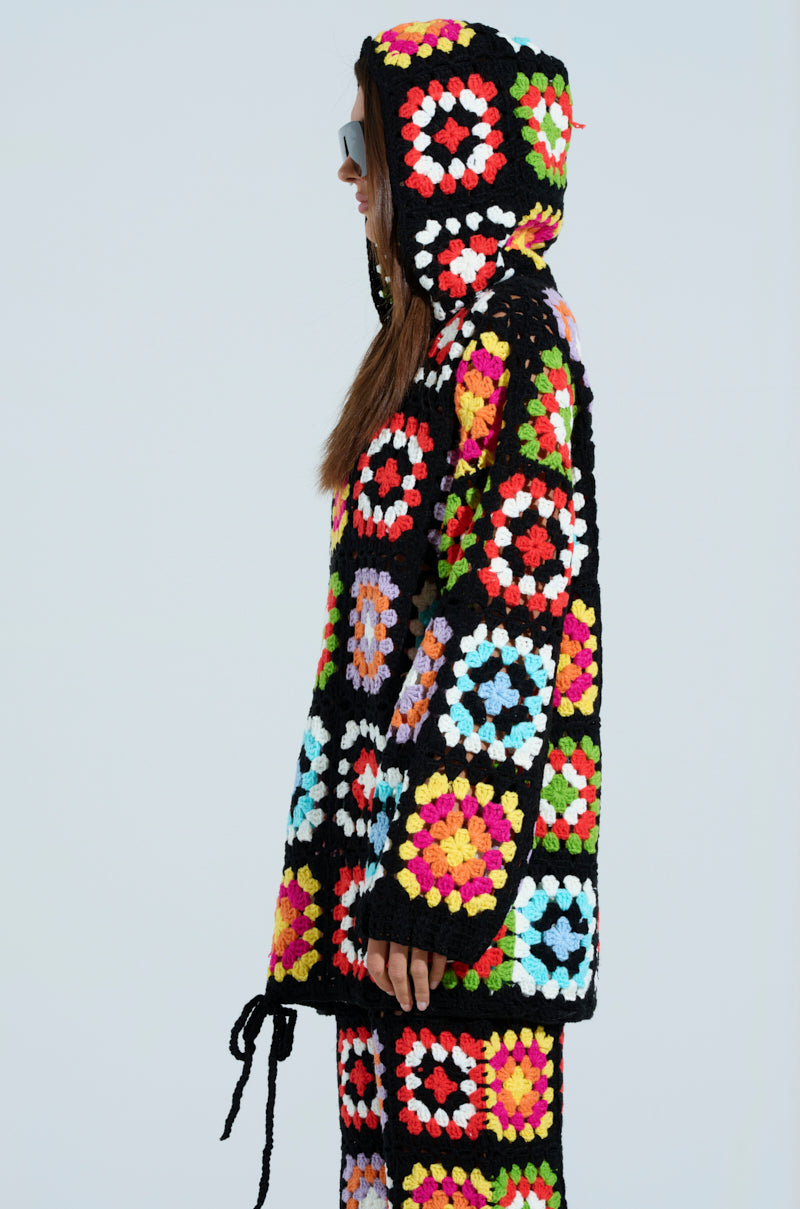 Wearing the Festival Girly Oversized Crochet Sweatshirt, this person dons a vibrant black hooded crochet outfit featuring multicolored squares. The ensemble includes matching pants, styled in an oversized silhouette with long hair flowing beneath the hood as they stand sideways.
