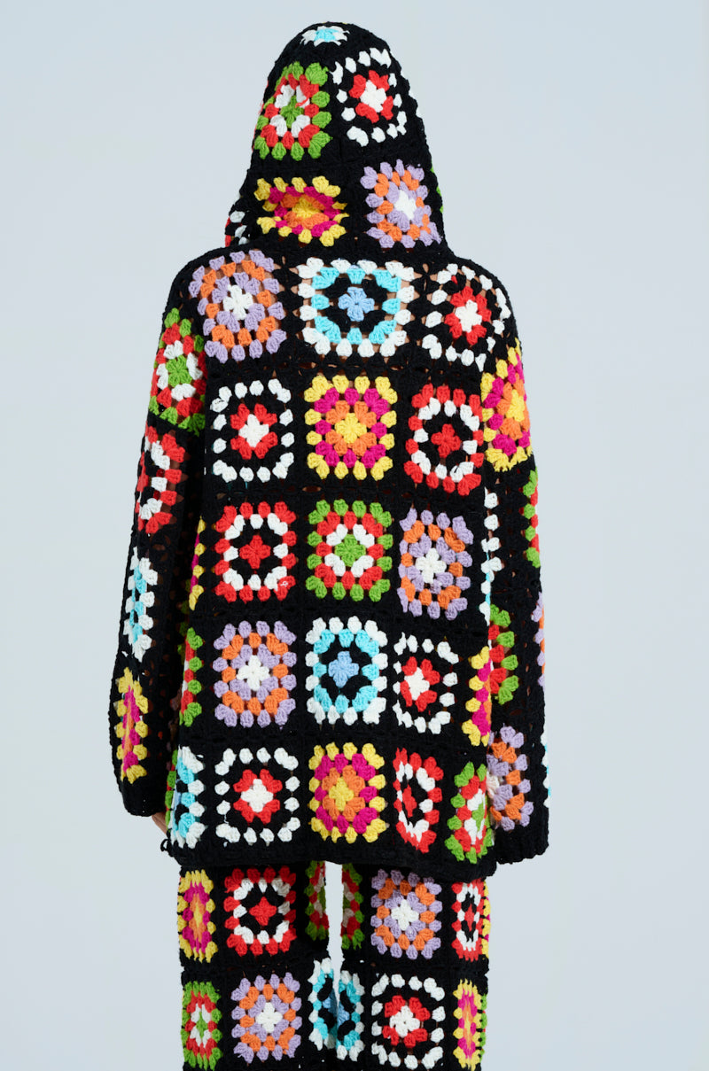 Against a gray background, a person wears the FESTIVAL GIRLY OVERSIZED CROCHET SWEATSHIRT by AKIRA Label, featuring a hooded, oversized design with a black base and multicolored square patterns, showcasing the unique and vibrant style.