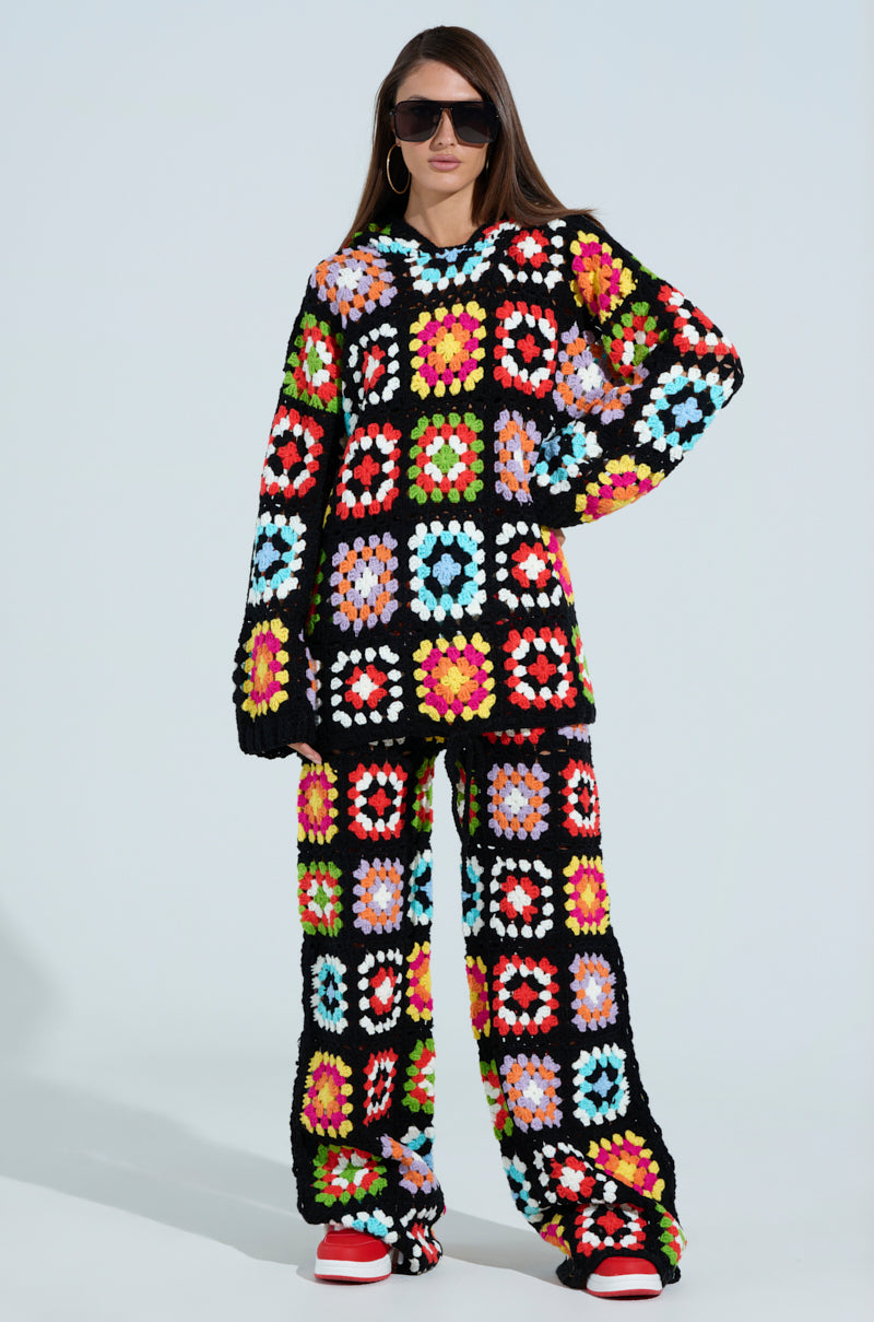 Dressed in a colorful AKIRA Label Festival Girly Oversized Crochet Sweatshirt, featuring a vibrant grid of multicolored squares, long sleeves, and an oversized silhouette, they pair it with wide-legged pants and red shoes. Large sunglasses frame their face as brown hair flows against the plain backdrop.