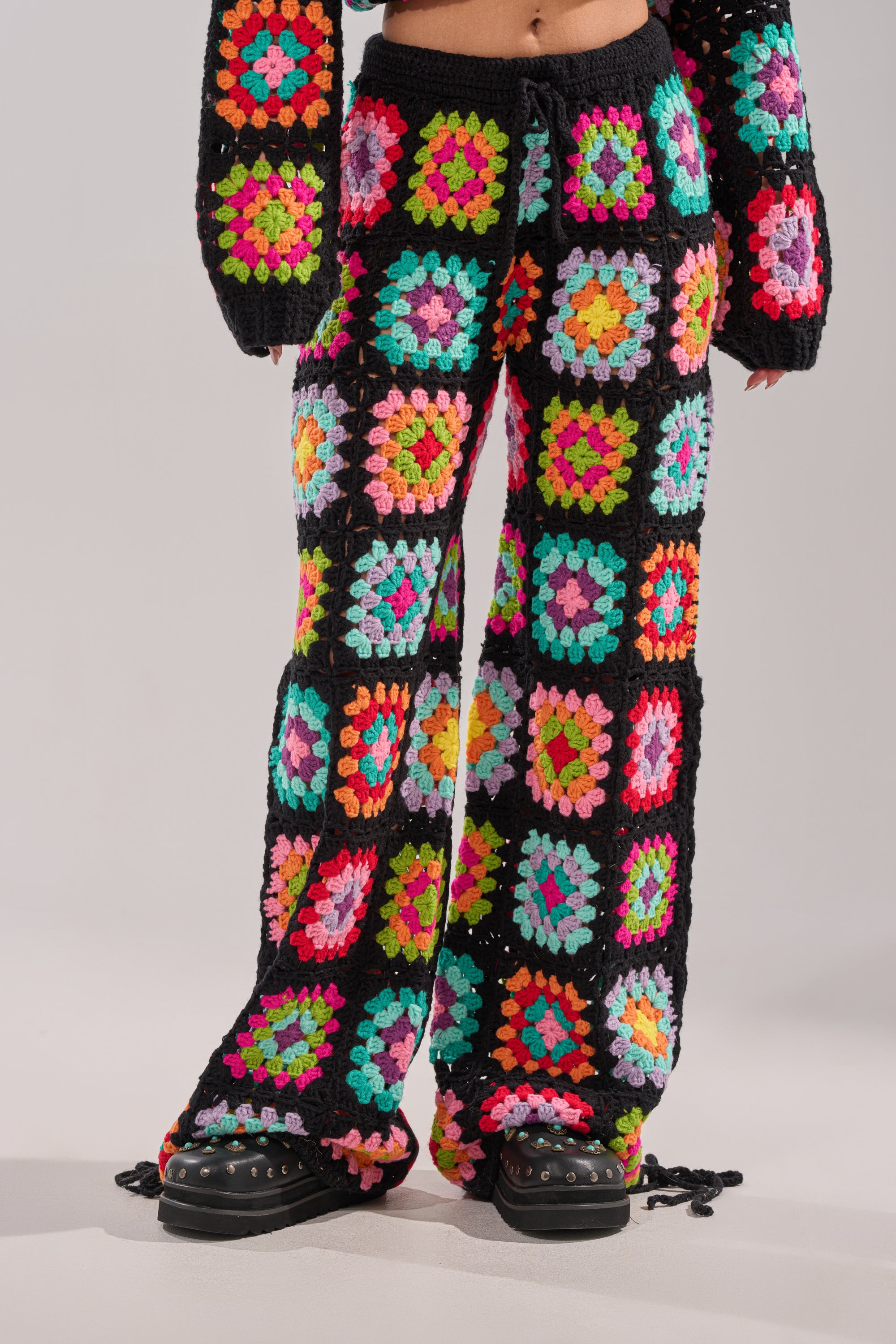 Wearing the FESTIVAL GIRLY OVERSIZED CROCHET PANT IN MULTI, featuring vibrant granny-square patchwork on a black base with matching sleeves—perfect for festival layering. Black shoes complete the colorful look. 0
