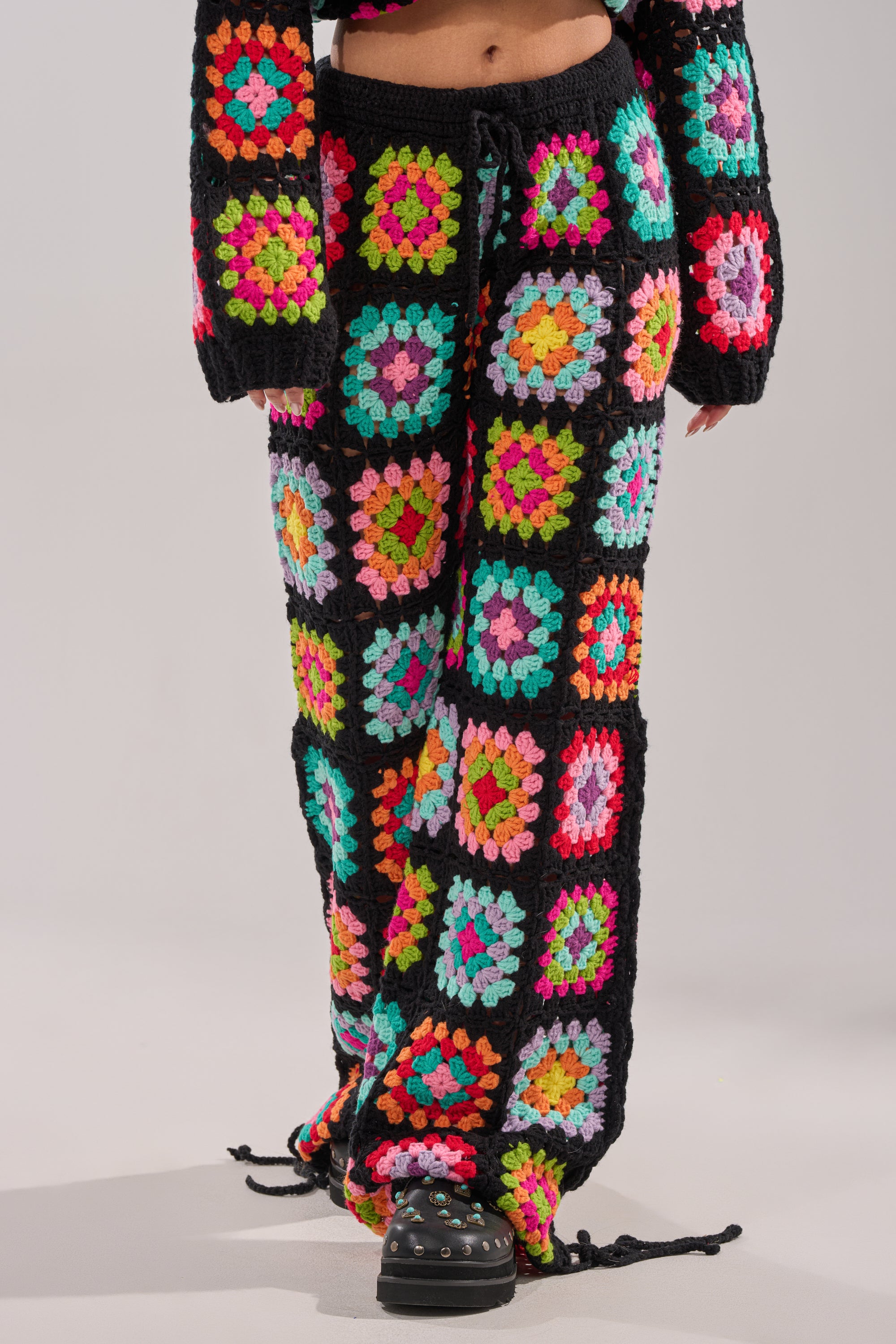 Wearing the FESTIVAL GIRLY OVERSIZED CROCHET PANT IN MULTI, featuring granny-square patchwork, wide legs, and a drawstring waist, styled with a matching long-sleeve top and black shoes—an ideal look for festival layering. 1