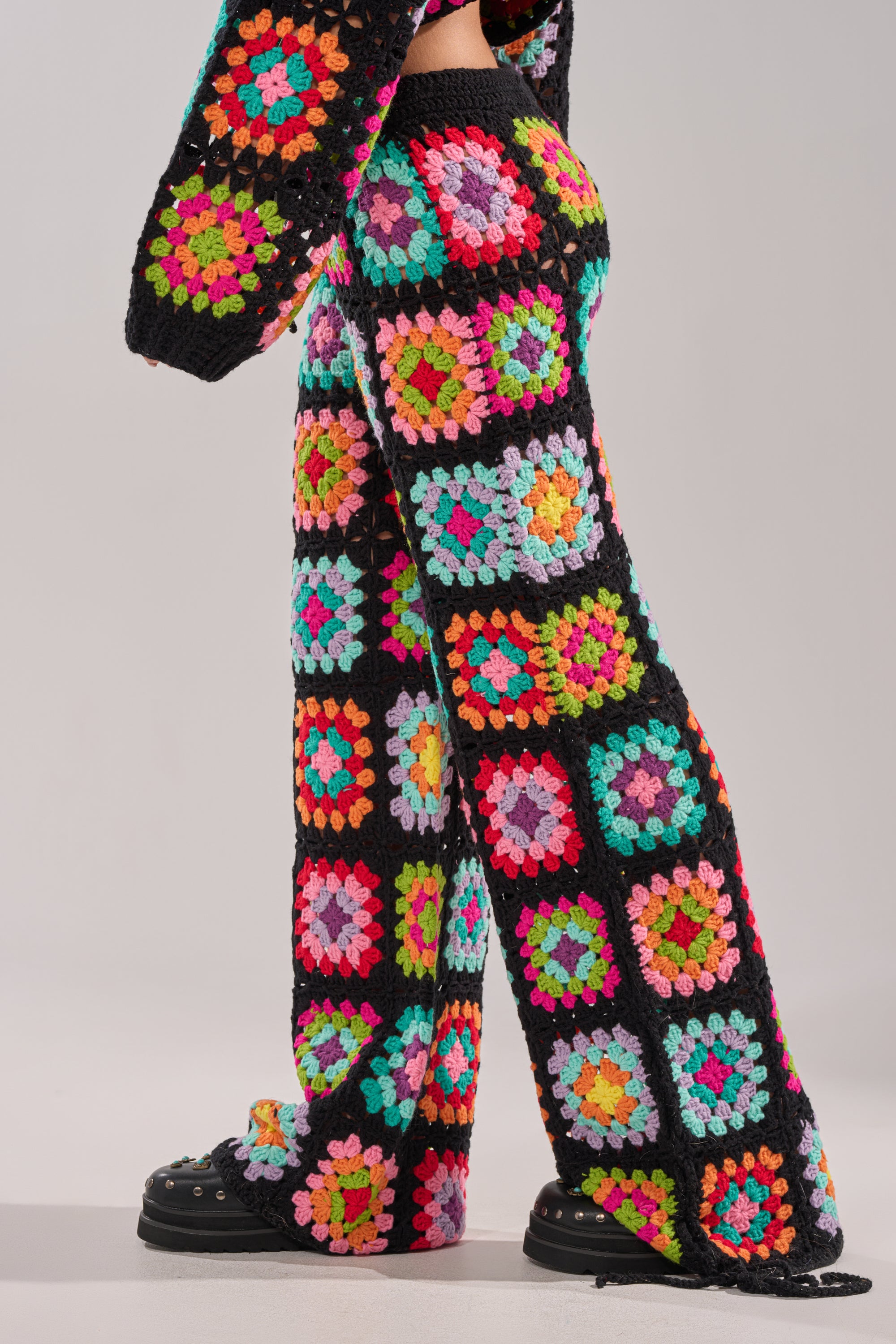 Model wears the FESTIVAL GIRLY OVERSIZED CROCHET PANT IN MULTI, featuring colorful granny-square patchwork and black outlines, styled with a matching long-sleeve top and black platform shoes against a plain light background. 2