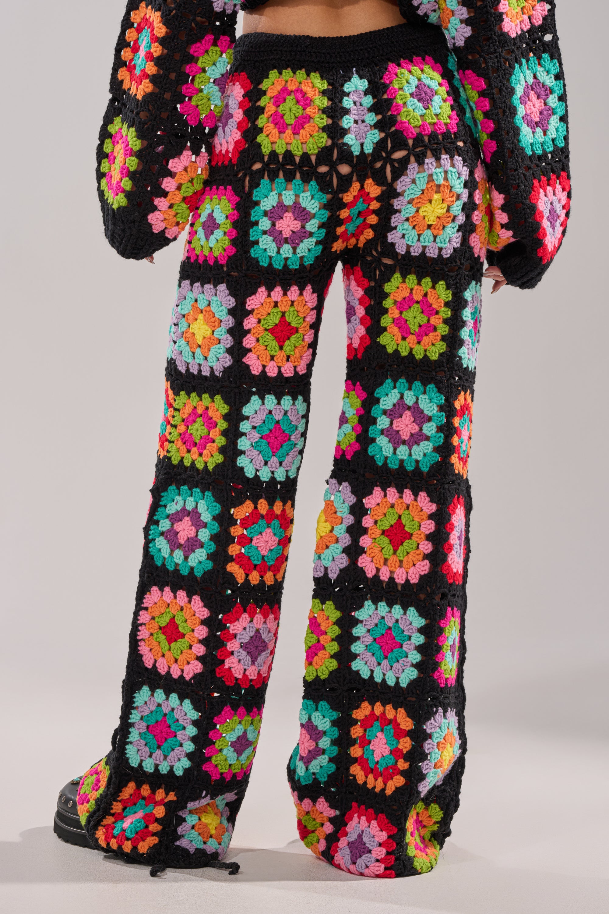 Someone wearing the FESTIVAL GIRLY OVERSIZED CROCHET PANT IN MULTI, featuring a vibrant multicolored granny-square patchwork design on a black background, is shown from behind. 3