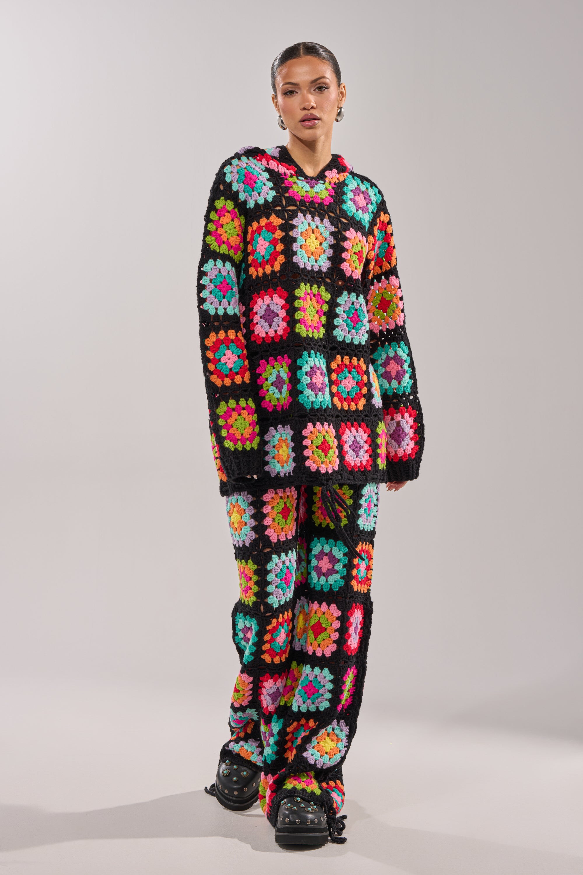 A person wears the FESTIVAL GIRLY OVERSIZED CROCHET PANT IN MULTI, a vibrant crochet knit set with pink, green, blue, orange, and yellow granny-square patchwork—ideal for festival layering with chunky black shoes. 5