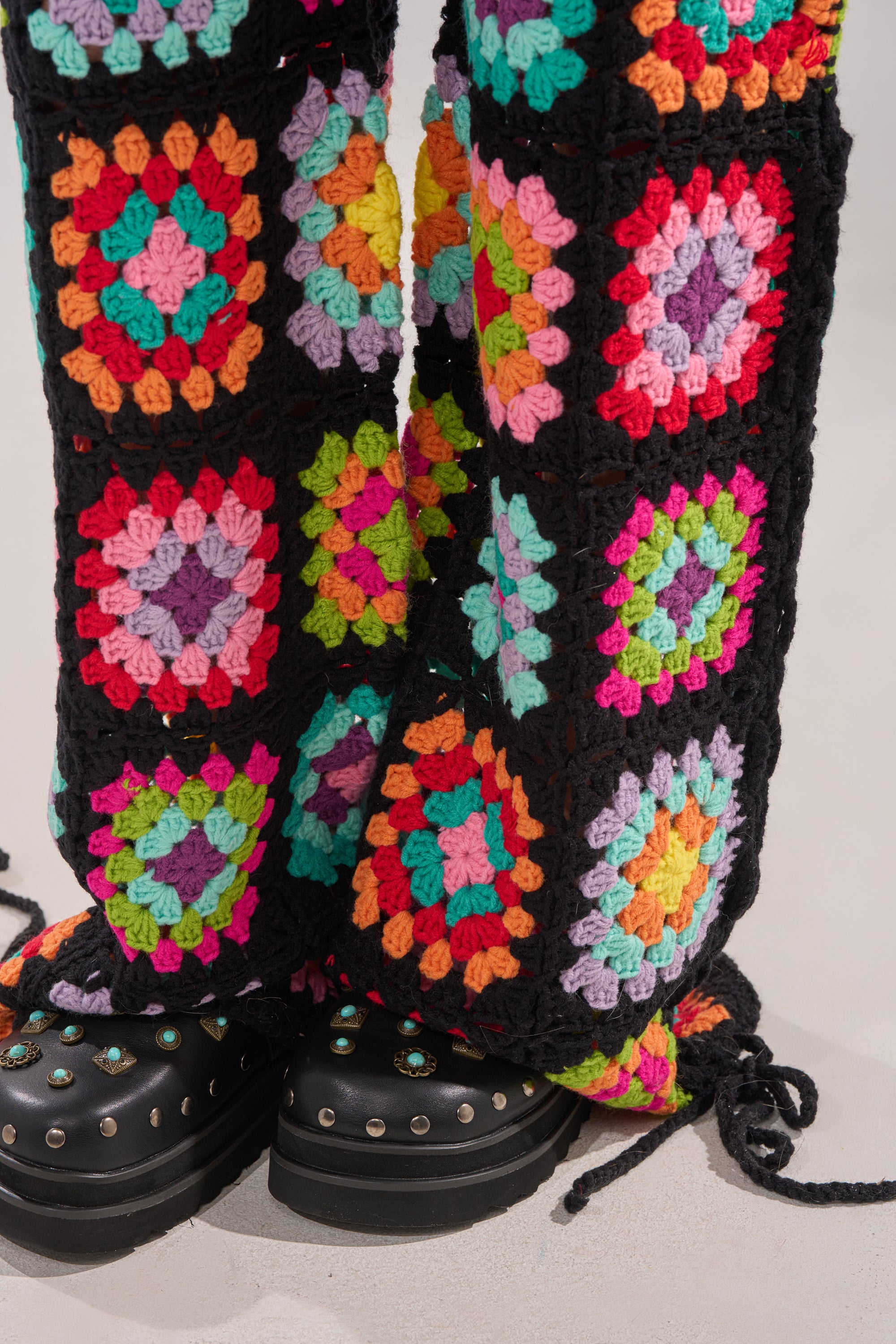 Close-up of legs in the FESTIVAL GIRLY OVERSIZED CROCHET PANT IN MULTI, showcasing vibrant granny-square knitwork, styled with black platform shoes trimmed with silver studs and turquoise, all set against a soft, neutral background. 7