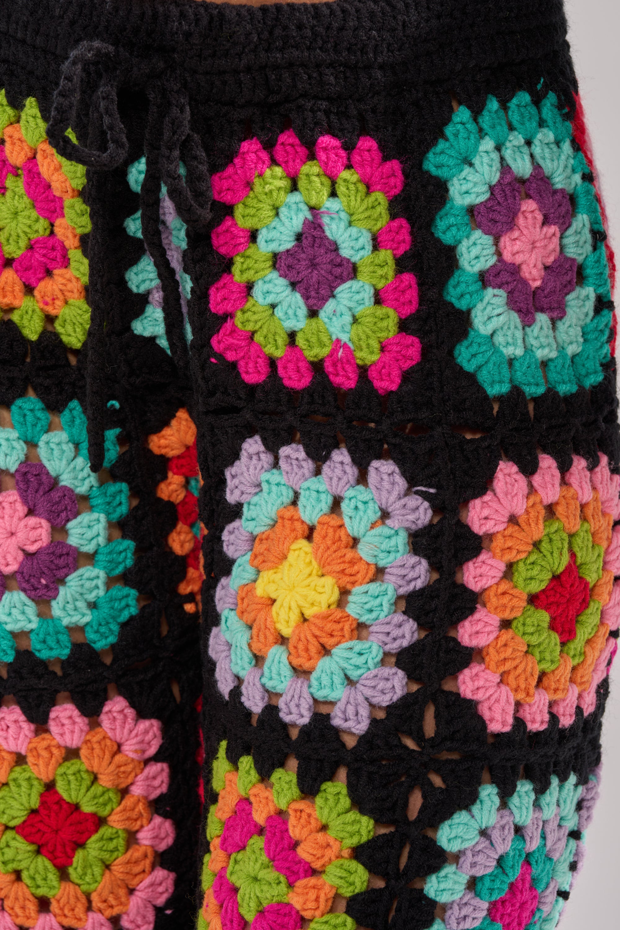 Close-up of the FESTIVAL GIRLY OVERSIZED CROCHET PANT IN MULTI, featuring bold granny-square patchwork in pink, green, orange, purple, yellow, and blue with black borders and a drawstring waist—ideal for festival layering. 8