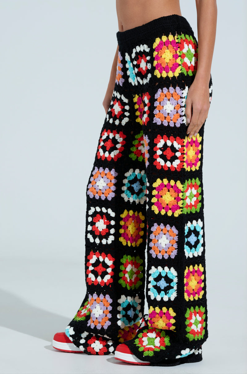A person wearing FESTIVAL GIRLY OVERSIZED CROCHET PANT with colorful granny square patterns and red shoes stands against a plain background.