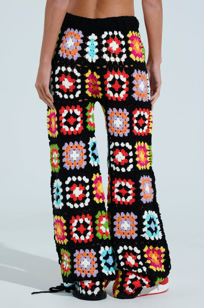 A person models the FESTIVAL GIRLY OVERSIZED CROCHET PANT boasting a vibrant granny square pattern in reds, pinks, greens, and blues. These AKIRA Label pants have a flared design that stands out against a neutral backdrop.