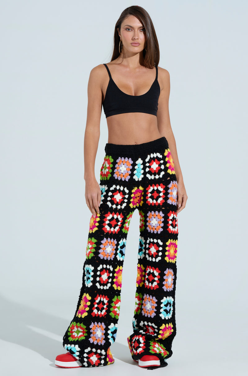 A woman in vibrant FESTIVAL GIRLY OVERSIZED CROCHET PANT from the AKIRA Label stands against a plain background. Her ensemble includes a black crop top and floral patterns, with long flowing hair and chic red and white sneakers, embodying a festival vibe.