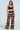 A woman in vibrant FESTIVAL GIRLY OVERSIZED CROCHET PANT from the AKIRA Label stands against a plain background. Her ensemble includes a black crop top and floral patterns, with long flowing hair and chic red and white sneakers, embodying a festival vibe.