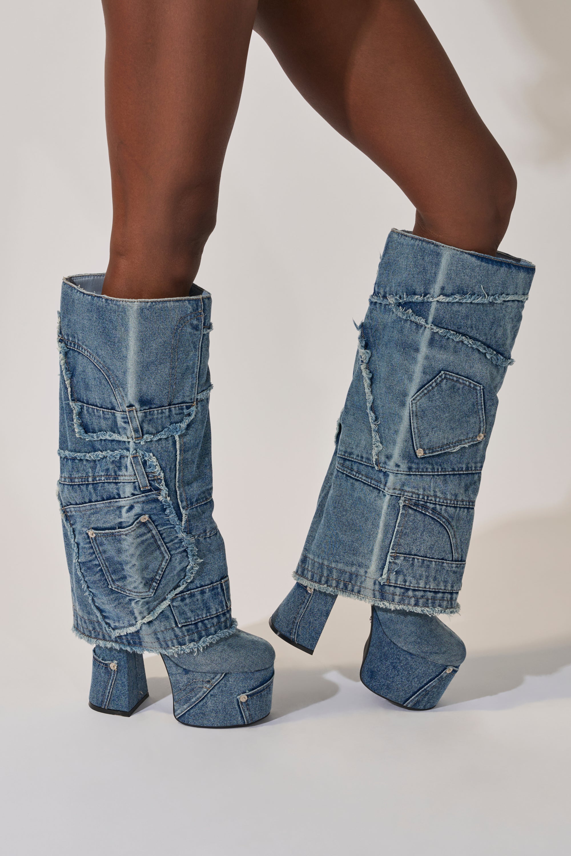 A person wears the AZALEA WANG DUMI DENIM CHUNKY PATCHWORK BOOT—featuring chunky platform soles, visible seams, frayed edges, and patchwork details—while standing against a plain light background. 2