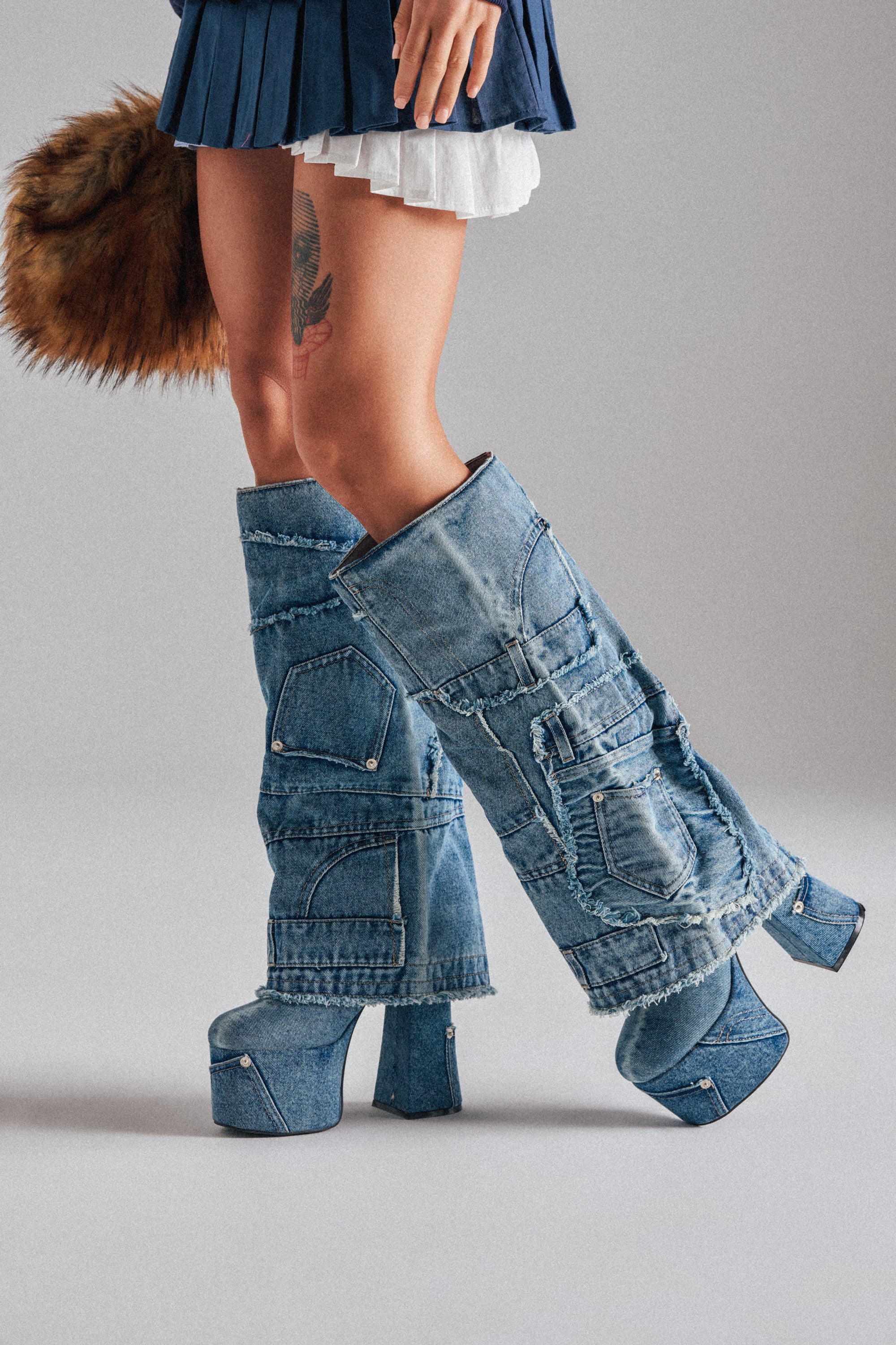 A person wearing the AZALEA WANG DUMI DENIM CHUNKY PATCHWORK BOOT, featuring a platform sole, blue jeans fabric with visible pockets and seams. They have a thigh tattoo and are dressed in a layered mini skirt. 0