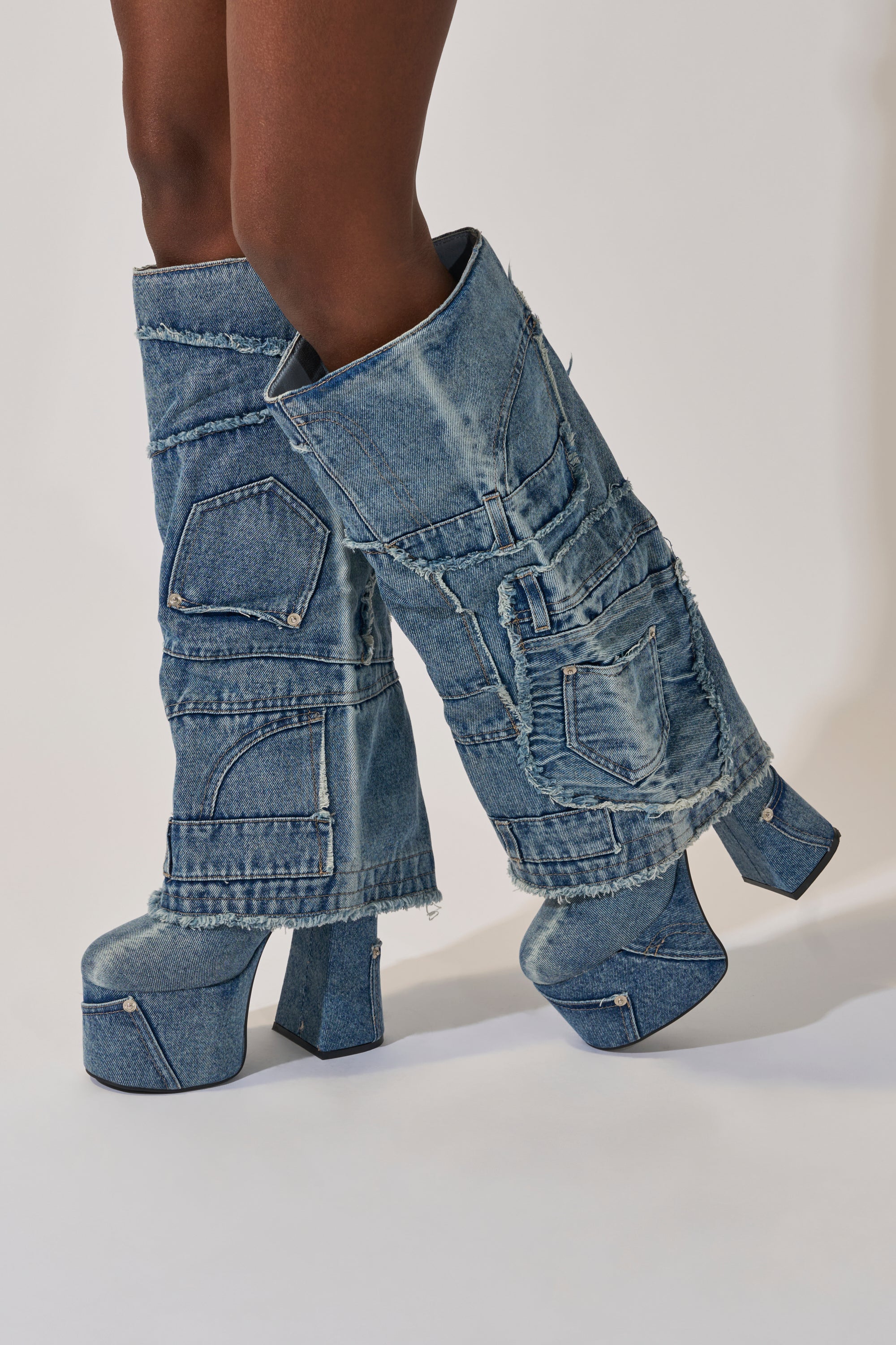 A person wears the AZALEA WANG DUMI DENIM CHUNKY PATCHWORK BOOT—knee-high with exposed seams, pocket details, and a bold platform sole—standing against a neutral background. 3