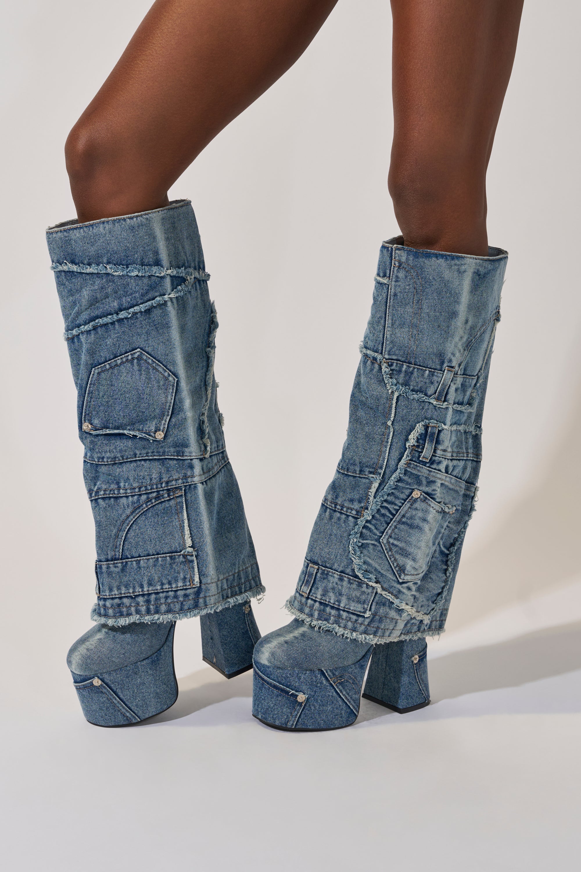 A person wears the AZALEA WANG DUMI DENIM CHUNKY PATCHWORK BOOT—knee-high footwear made from repurposed blue denim jeans with visible pockets, seams, and frayed edges—standing against a light background. 4