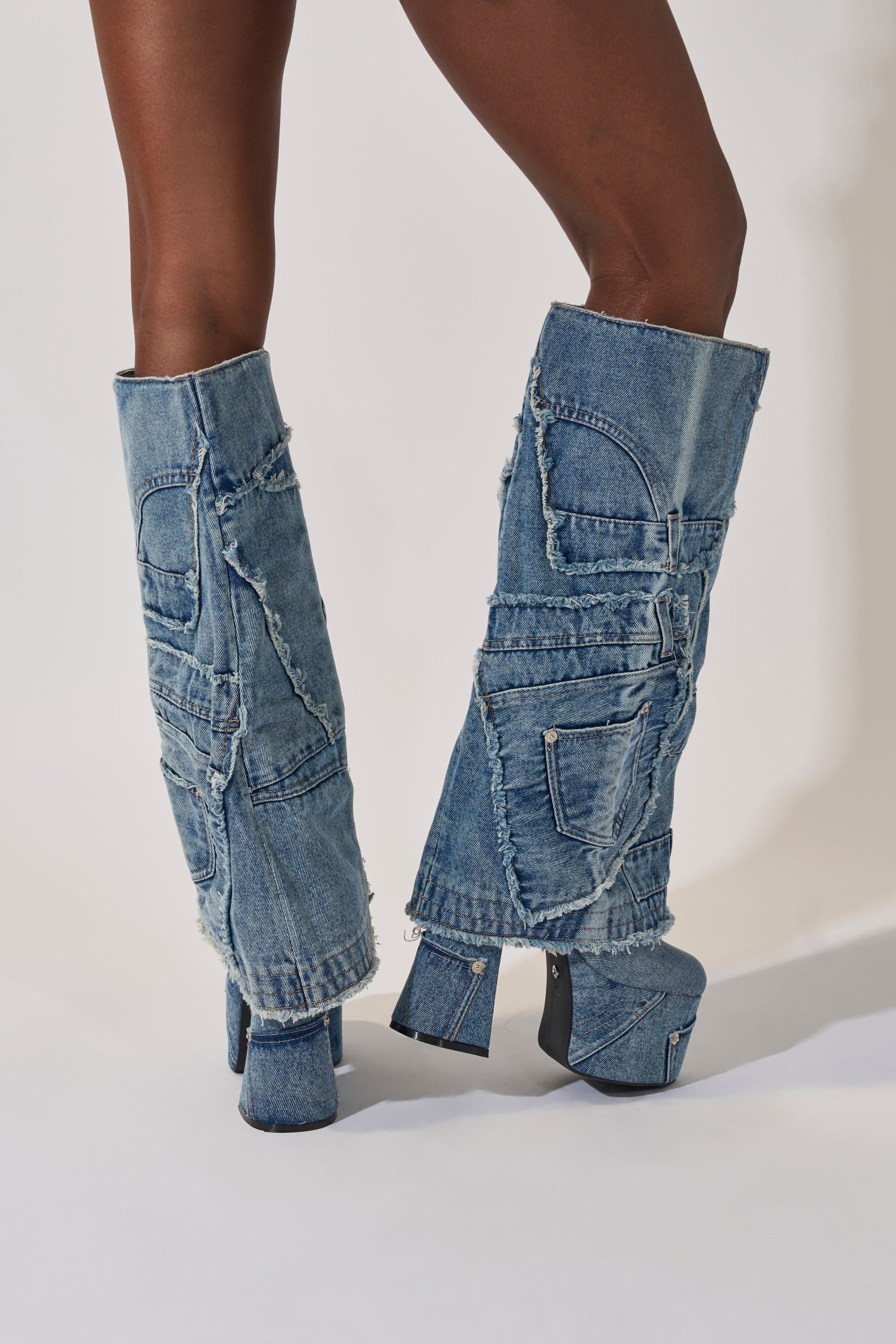 A person models the AZALEA WANG DUMI DENIM CHUNKY PATCHWORK BOOT, featuring knee-high patchwork denim, frayed edges, multiple jean pockets, and squared high heels against a plain background. 9