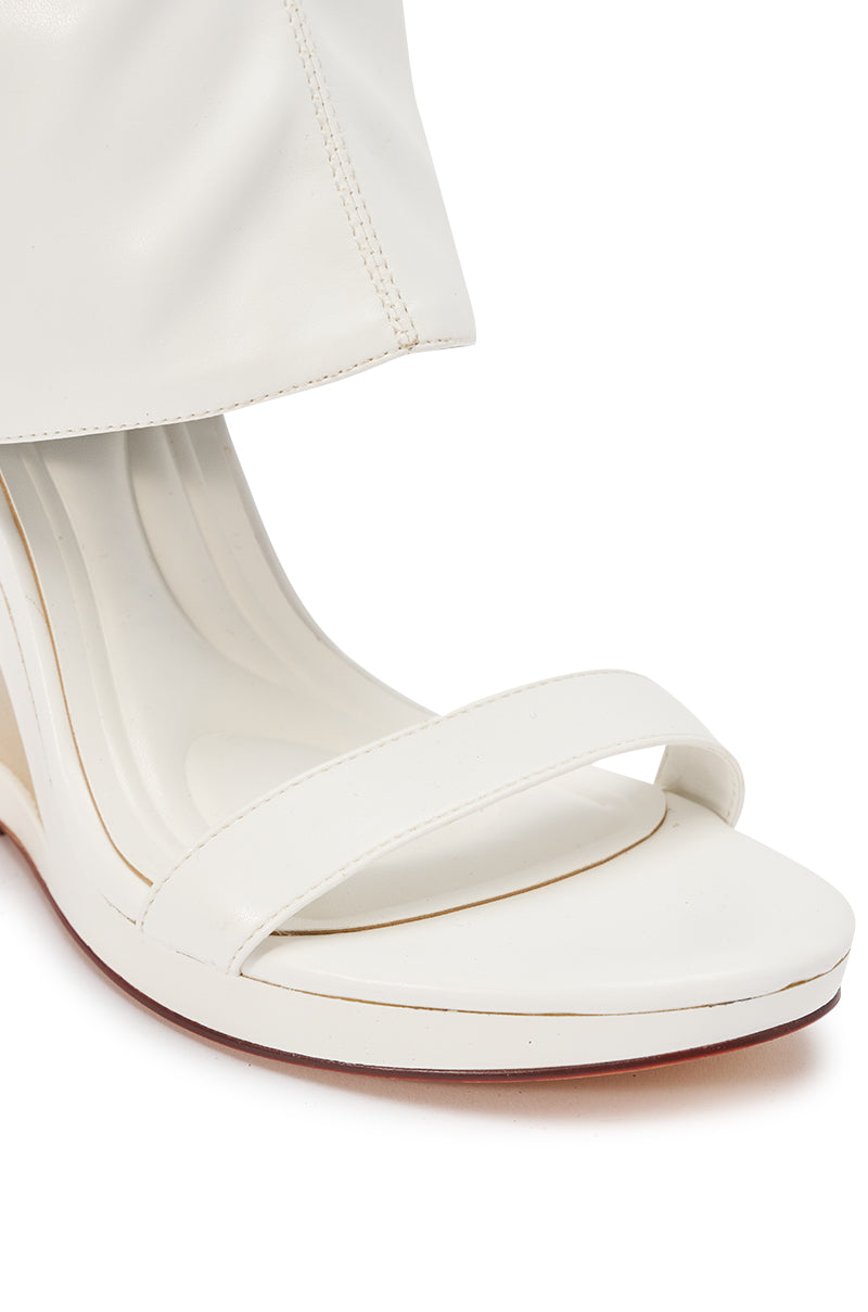 A close-up of the AKIRA GOLD DUMISANI WHITE WEDGE OPEN TOE BOOTIE, featuring a square toe and single strap, worn with white pants that partially cover the shoe.