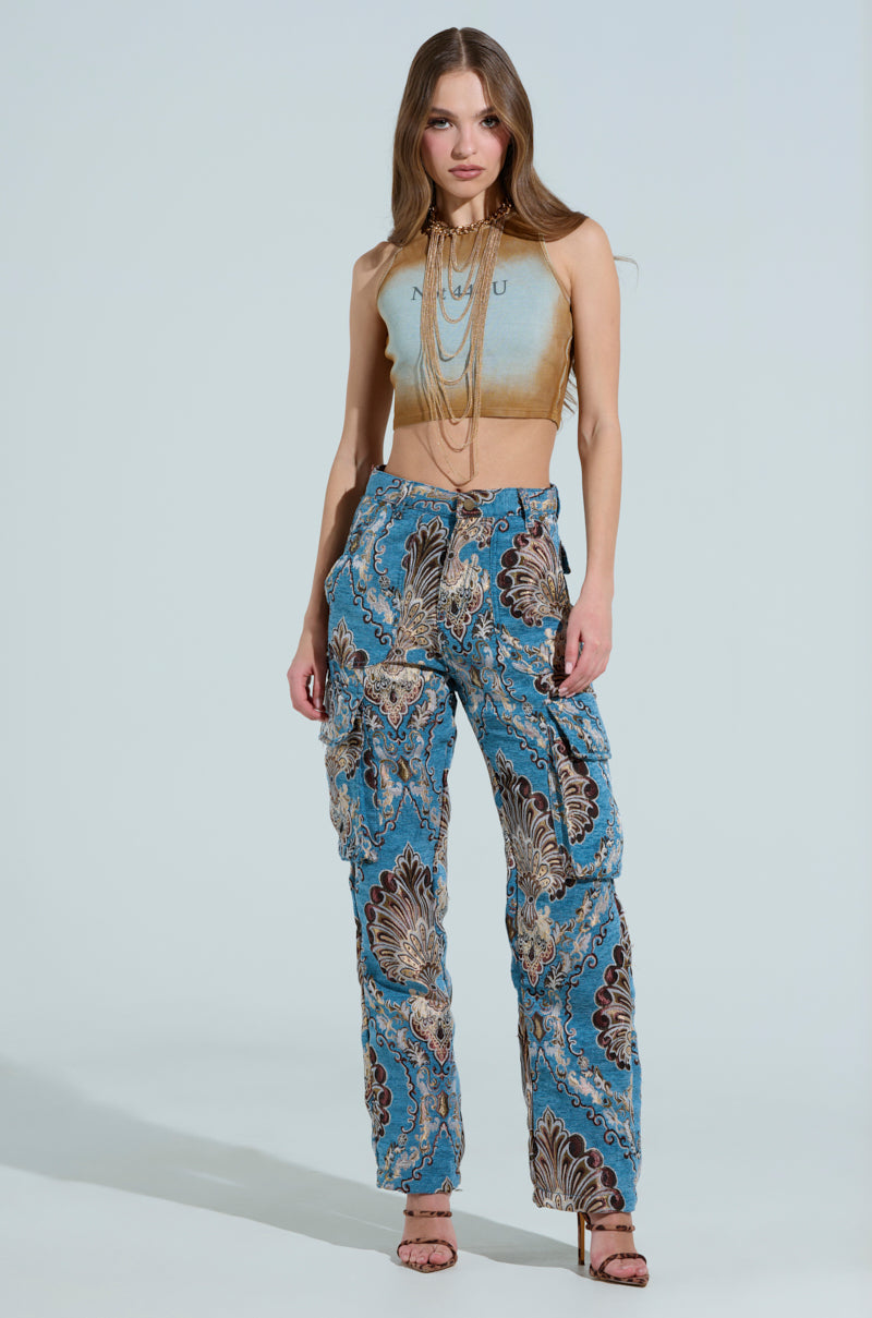 Confidently standing with hands in the cargo pockets of their THE DUOMO PANT, they sport a sleeveless light blue crop top, heeled sandals, and a beaded necklace against a plain background.