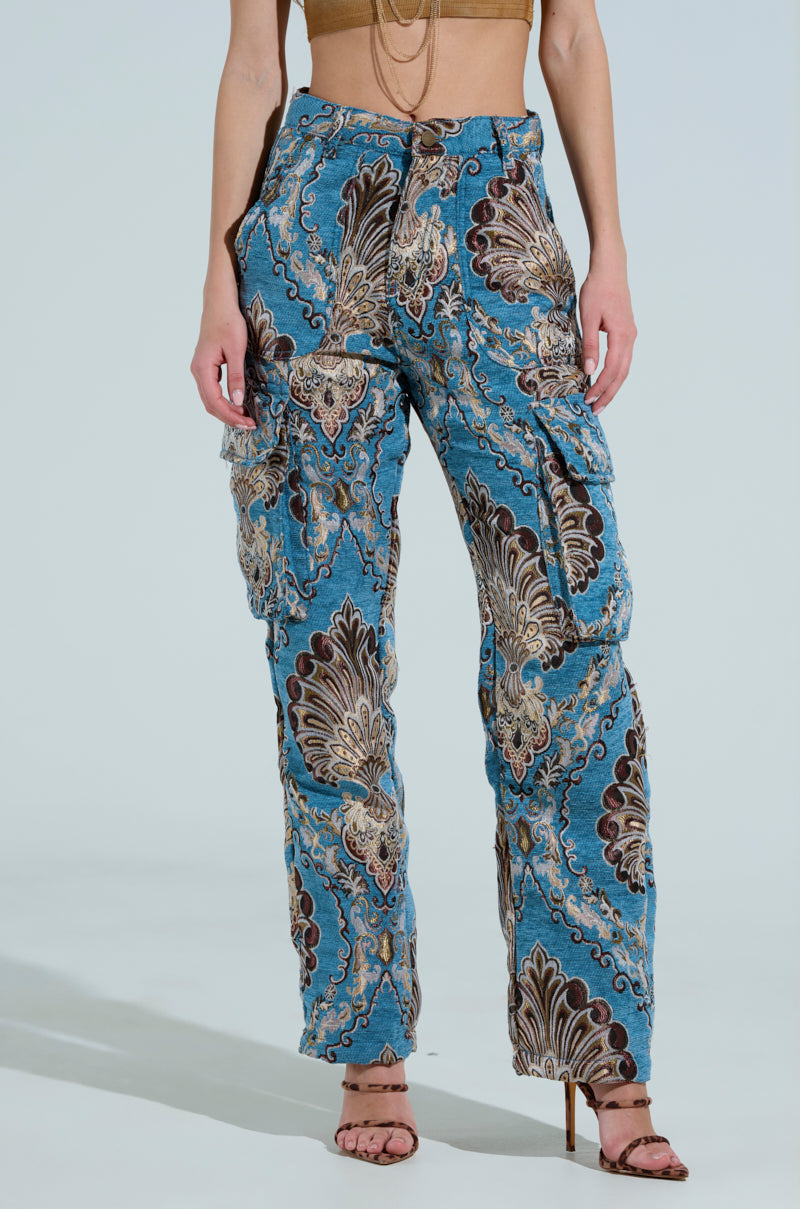 Someone wearing THE DUOMO PANT in blue suede-like jacquard with a paisley pattern stands against a light background. Paired with a tan crop top and open-toed heels, the high-rise pants feature multiple cargo pockets and intricate designs, enhancing their stylish appeal.