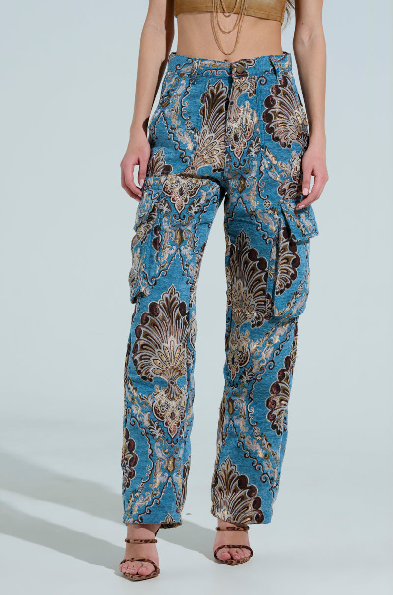 A woman models THE DUOMO PANT, featuring wide-legged high-rise design in a blue and brown paisley pattern, paired with a beige cropped top adorned with a chain. She poses against a light blue background, completing the outfit with strappy heels.
