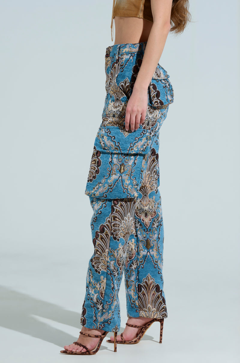 A person wearing a brown crop top and THE DUOMO PANT, which are stylish blue cargo pants with paisley designs, stands against a neutral background. The high-rise pants have cargo pockets and are perfectly paired with open-toed high heels, capturing a chic side view.