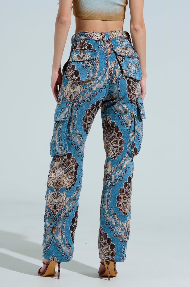 A person showcases THE DUOMO PANT, a pair of suede-like jacquard cargo pants with large pockets and a blue and brown paisley pattern, paired with a light-colored cropped top. They stand with their back facing, highlighting high-heeled sandals against a plain light blue background.