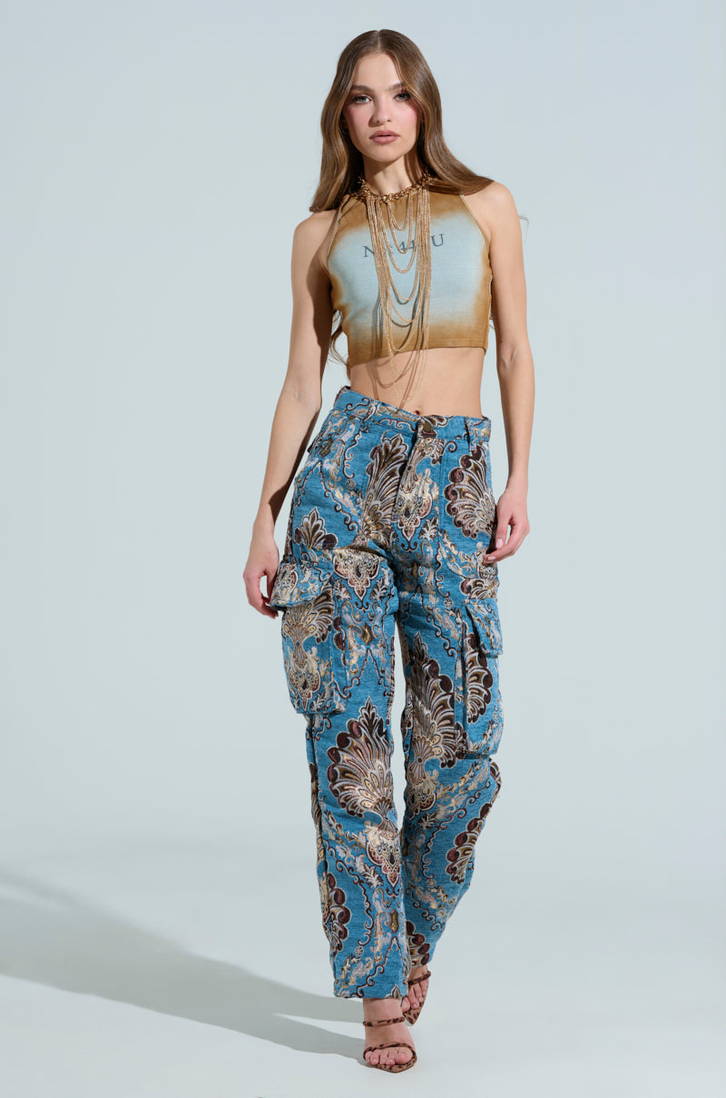 A person with long hair walks in a sleeveless crop top and THE DUOMO PANT, suede-like jacquard cargo pants in shades of blue and brown. The high-rise pants are perfectly paired with strappy high-heeled sandals against a light grey background.