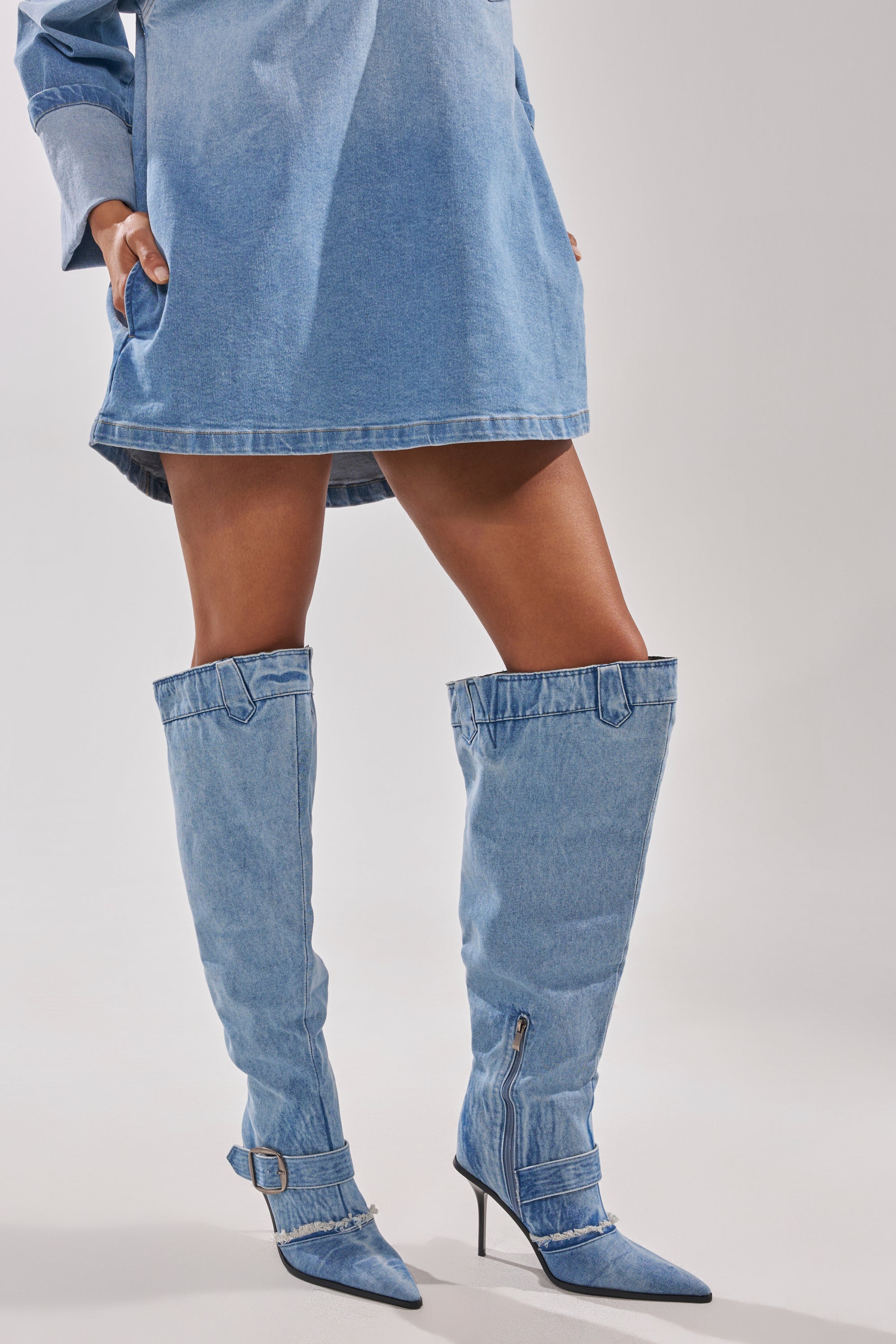 Someone stands against a light background wearing a blue denim dress and AKIRA GOLD DURANGO DENIM STILETTO PANT LOOK BOOTS featuring pointed toes, buckles, zippers, and an over-the-knee silhouette. 1