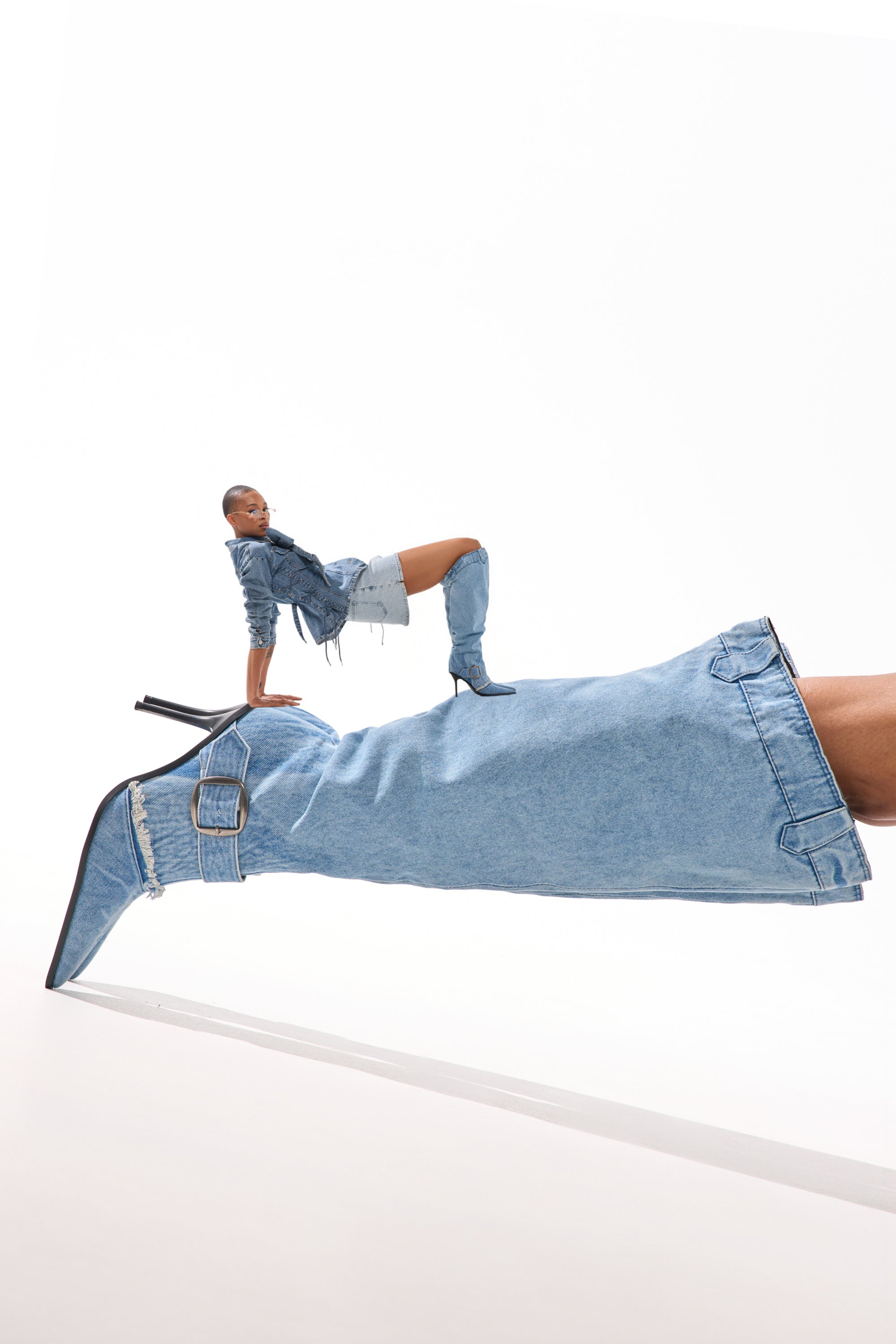 A woman models the AKIRA GOLD DURANGO DENIM STILETTO PANT LOOK BOOT, posing atop an oversized denim stiletto boot and creating a striking optical illusion against a white background. 0