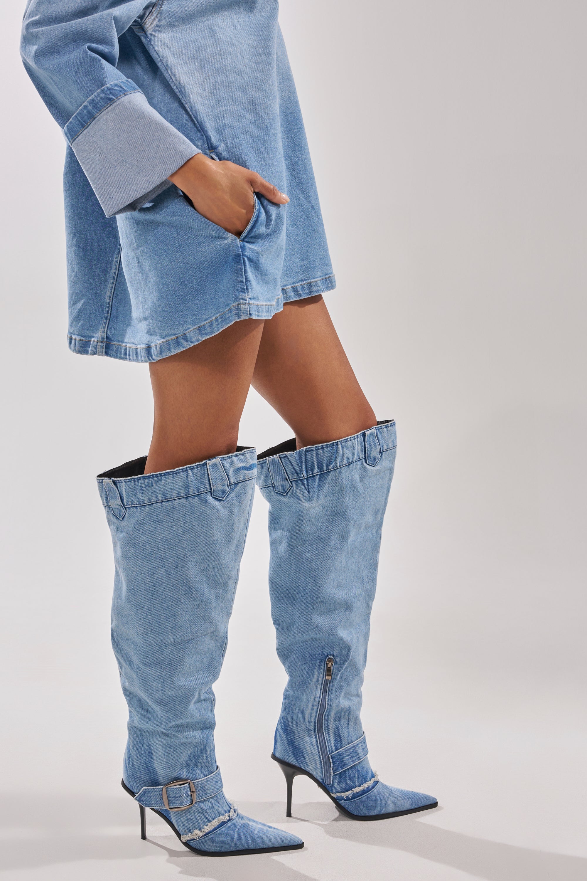 A person wears a blue denim dress and the AKIRA GOLD DURANGO DENIM STILETTO PANT LOOK BOOT—over-the-knee, pointed-toe boots with buckles—standing against a plain light background. 2
