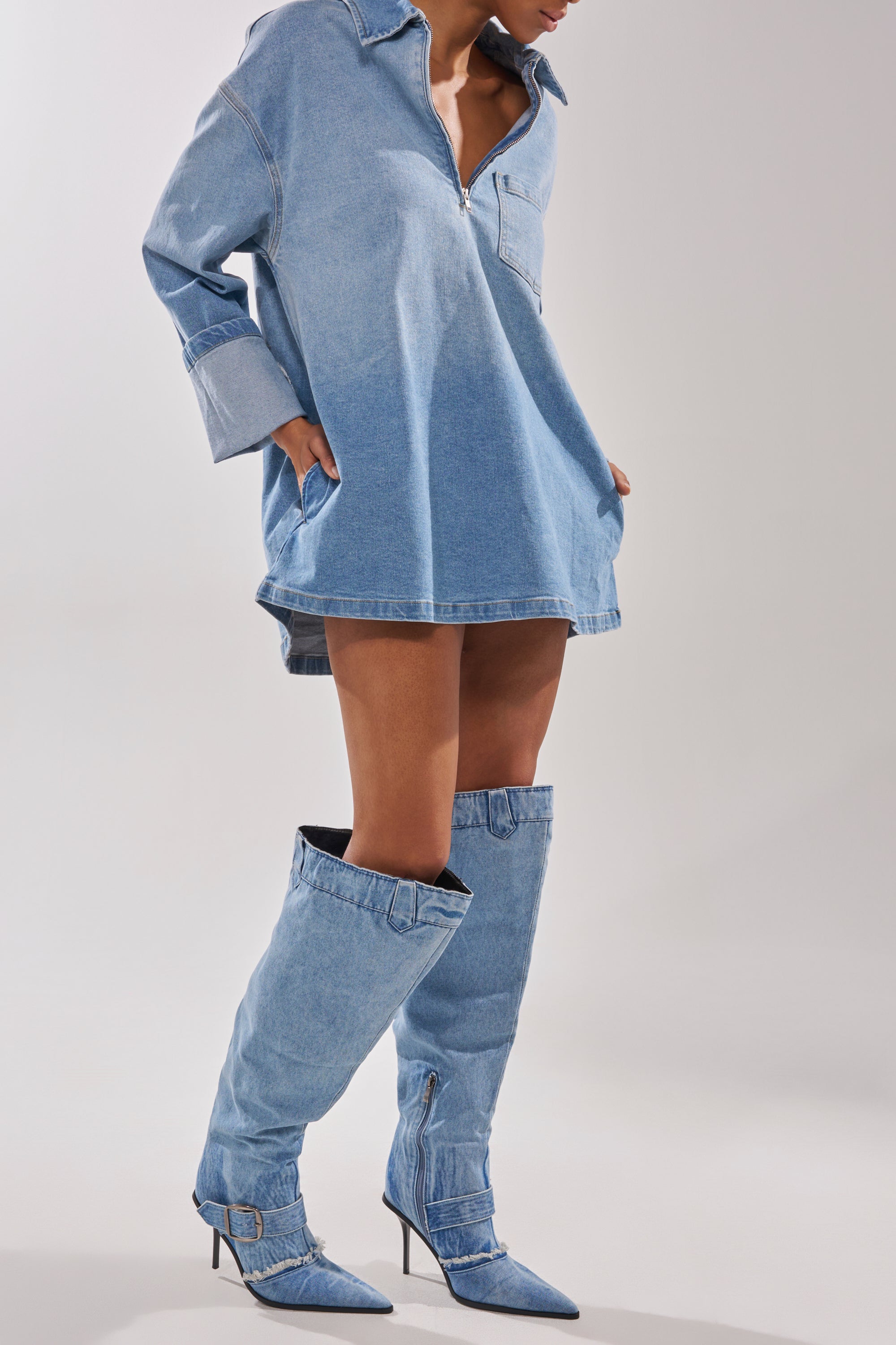 A person stands against a plain light background wearing a loose, long-sleeve denim shirt dress and the AKIRA GOLD DURANGO DENIM STILETTO PANT LOOK BOOT with pointed toes. 3