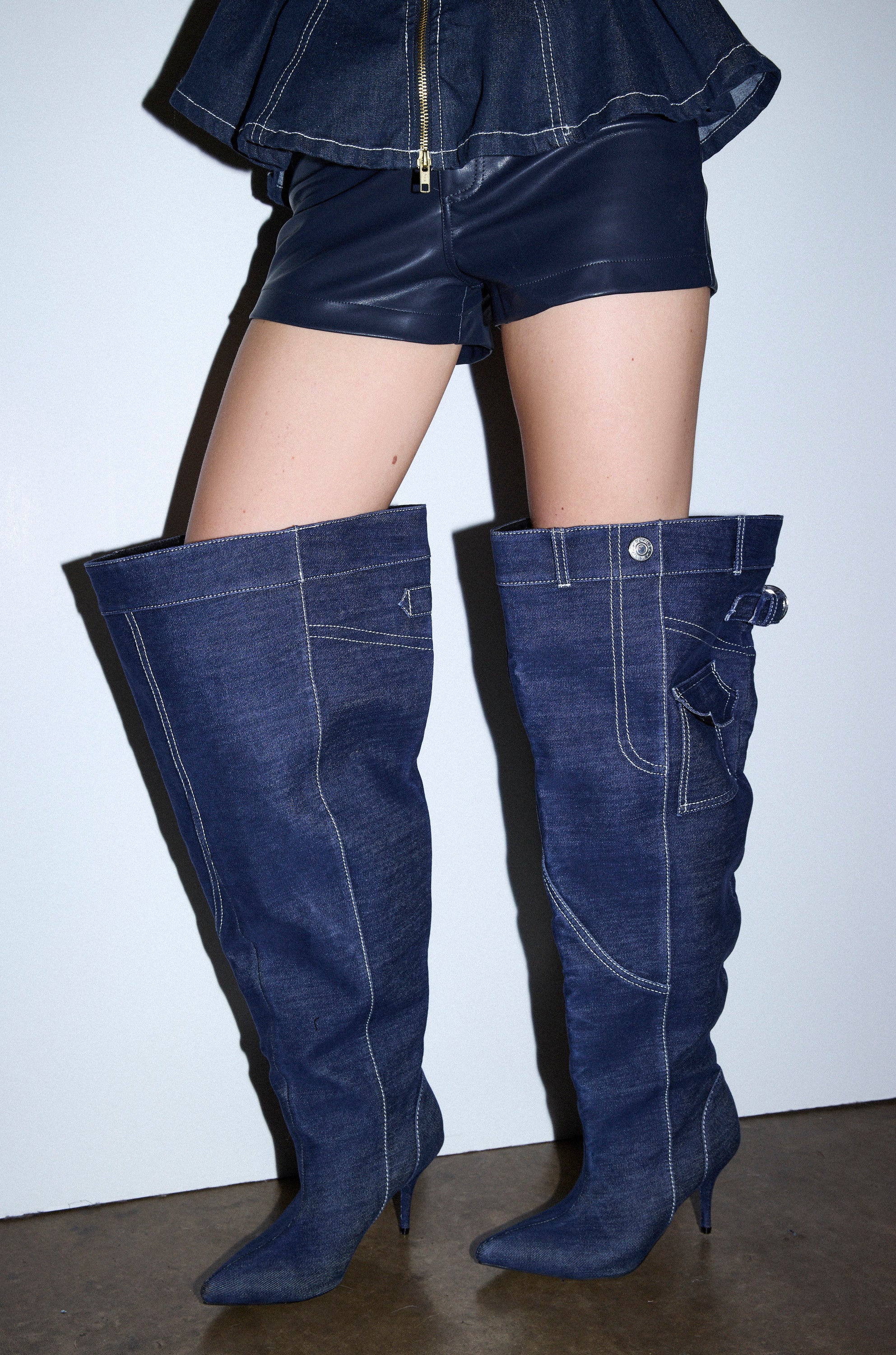 A person wears the AZALEA WANG DUSTAN DENIM THIGH STILETTO BOOT with pointed toes, styled with black leather shorts and a zip-up top, standing against a plain white background.