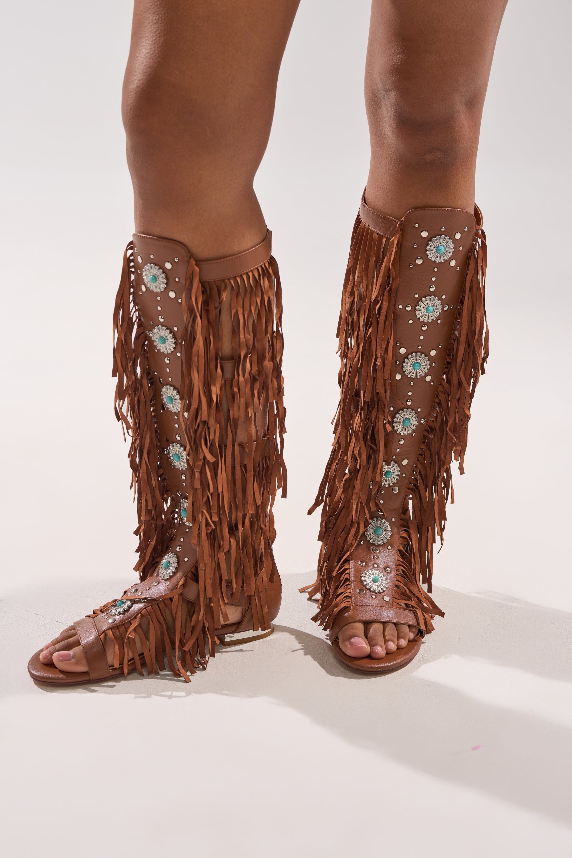 A person wears the AZALEA WANG DUSTINE BROWN GLADIATOR EMBELLISHED SANDAL, featuring knee-high straps with turquoise and silver details, while standing on a light background. 2