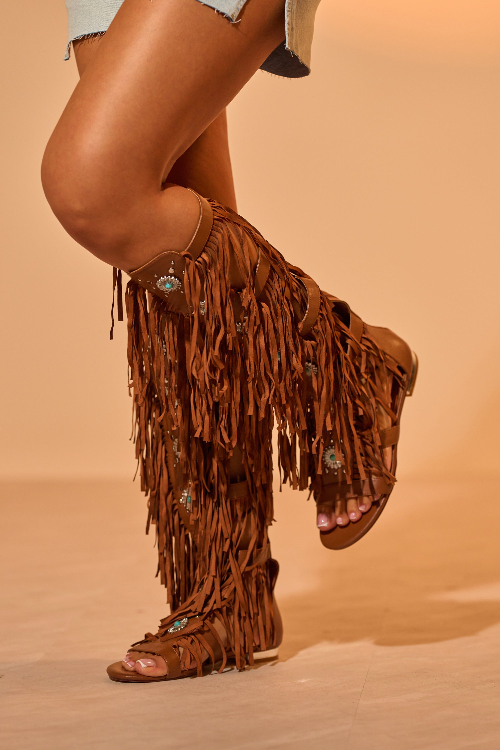 A person stands on a wooden floor in a light blue denim skirt, wearing AZALEA WANG DUSTINE BROWN GLADIATOR EMBELLISHED SANDALS with turquoise accents. The background is a soft tan color. 1