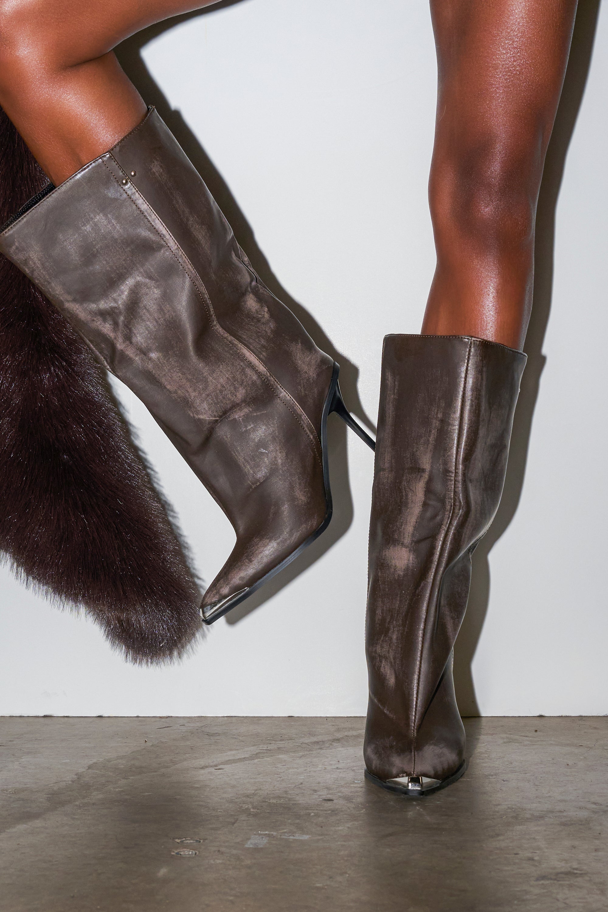 A person wears AZALEA WANG DUSTY BROWN WEATHERED PU BOOTS with stiletto heels, standing against a white wall on a concrete floor. A dark fur garment is partially visible at the left edge of the frame.