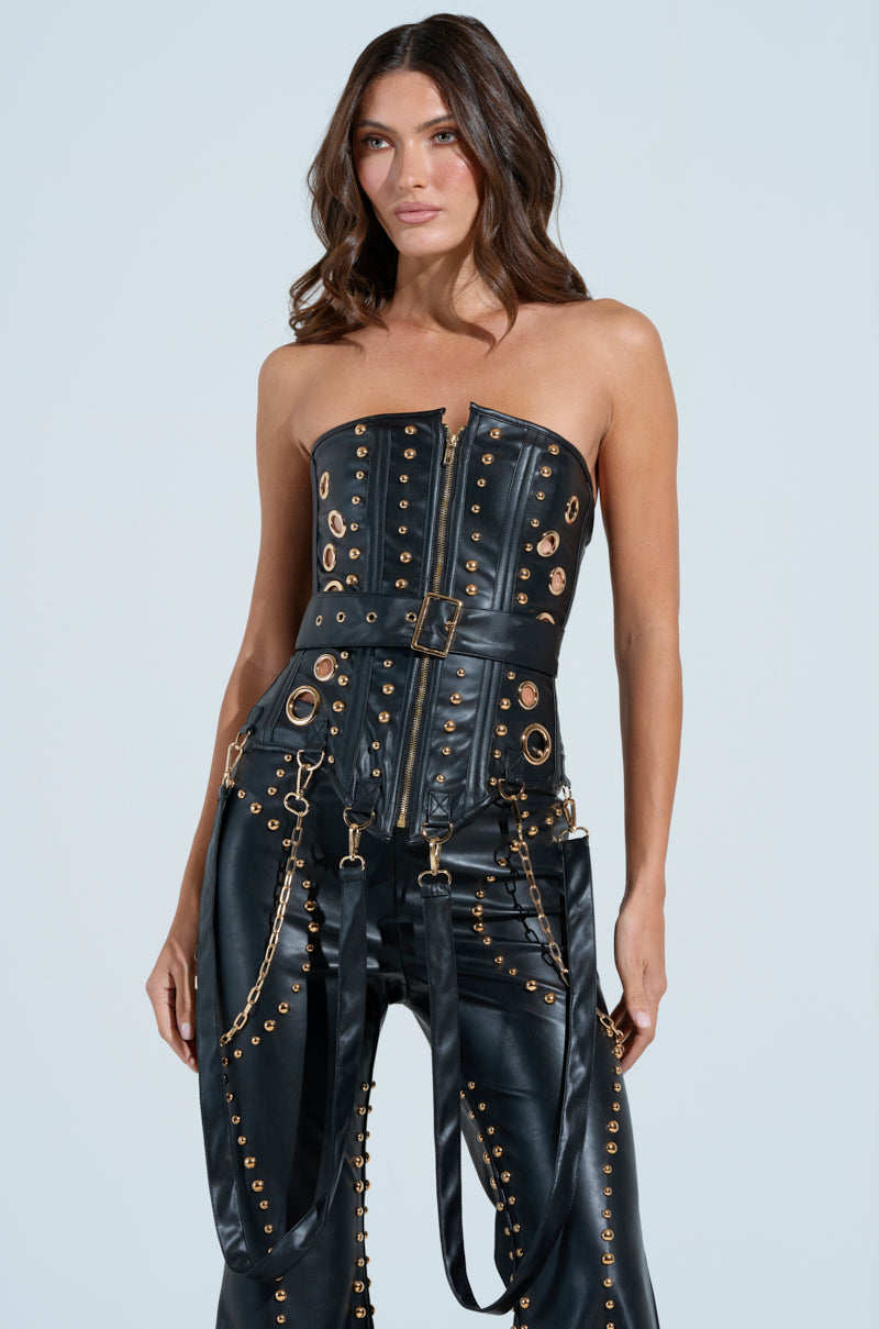 A woman poses against a light background in the DW STUDDED CORSET, featuring black faux leather with gold studs and metallic accents. The ensemble includes a belted top, matching pants with chain details, and her hair styled in loose waves.