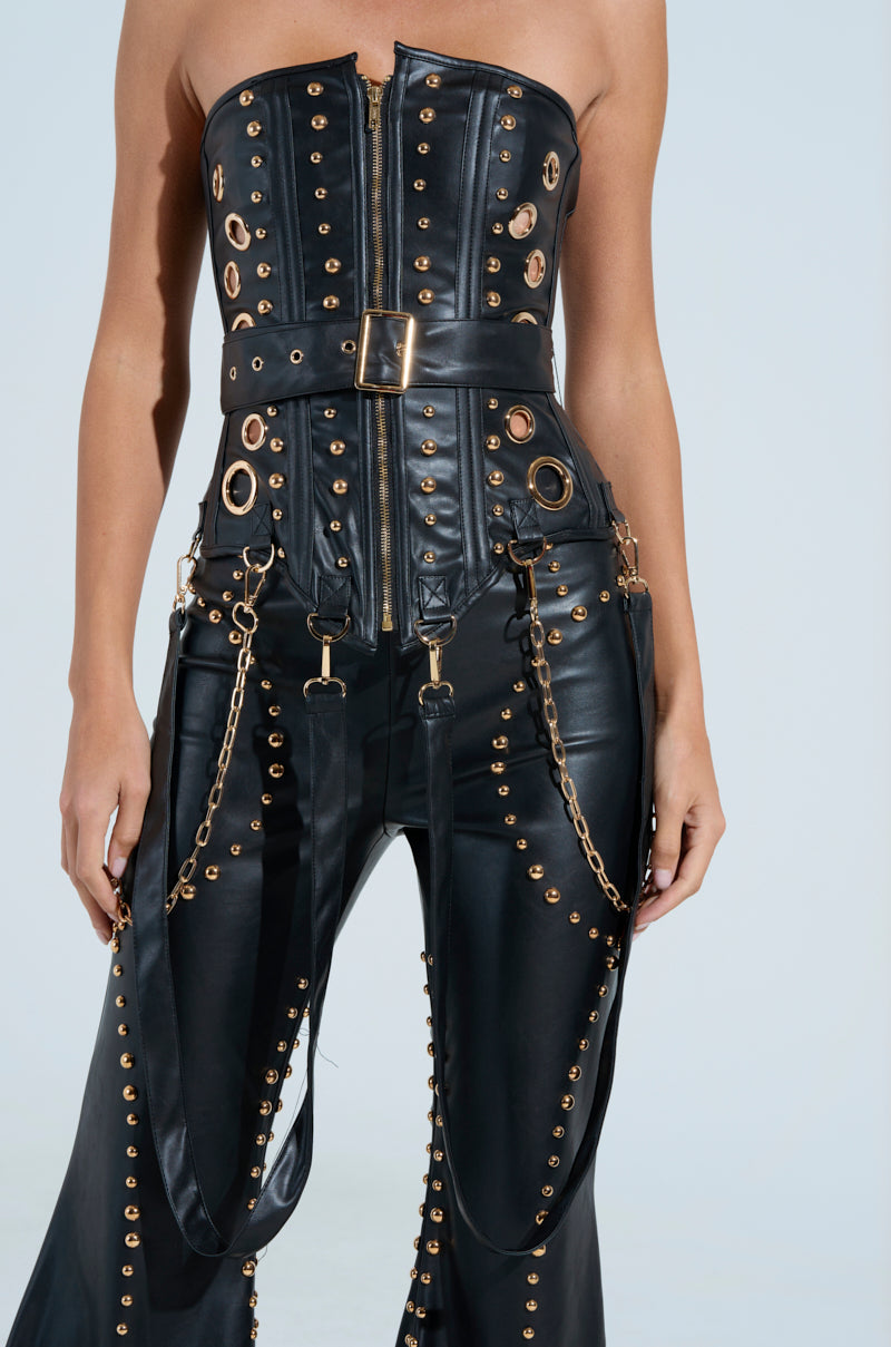 Wearing the DW STUDDED CORSET, a sleek black faux leather ensemble, highlighted by metallic hardware with eyelets, chains, and straps. Featuring a strapless top with a front zipper and matching pants against a plain light backdrop.