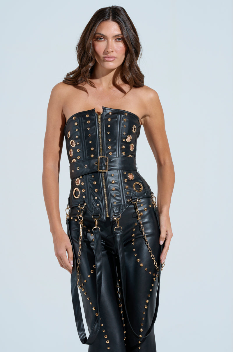 A woman in a stylish black outfit featuring metallic studs and chains stands against a plain backdrop. The ensemble includes a strapless DW STUDDED CORSET paired with matching pants, creating a bold fashion statement.